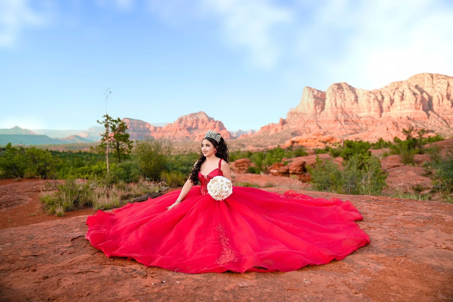 Young woman in a voluminous red lace ball gown poses with bouquet on red rocks in Sedona, Arizona.