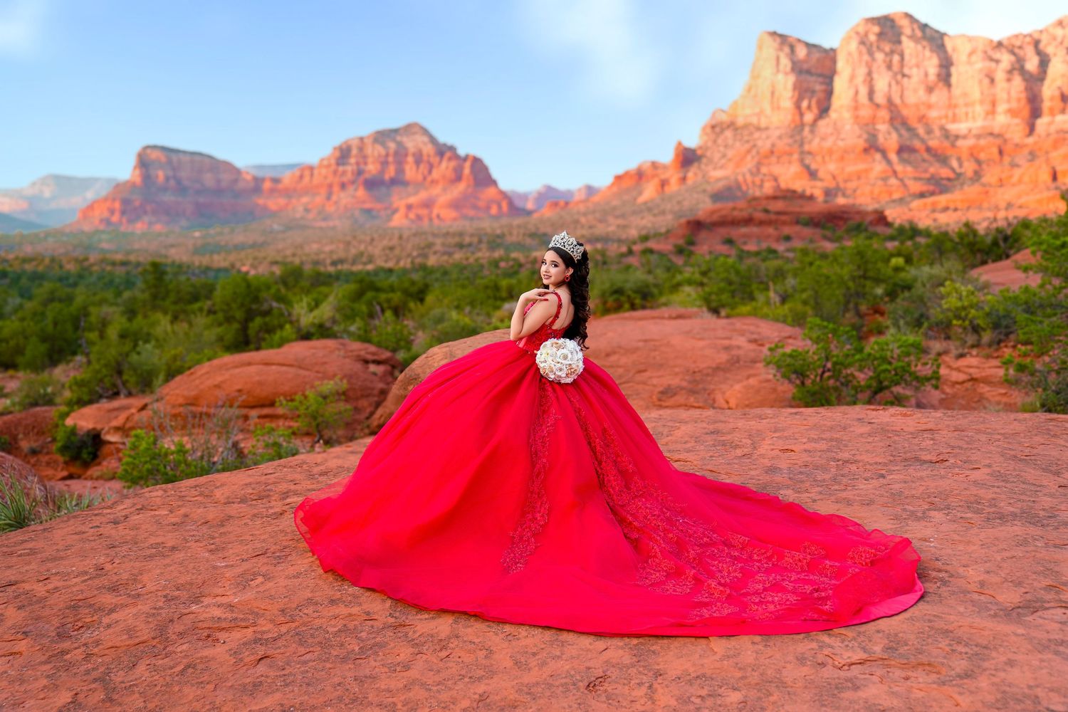 Woman in an elegant red quinceañera ball gown sits on red rocks with Sedona's scenic landscape behind her.