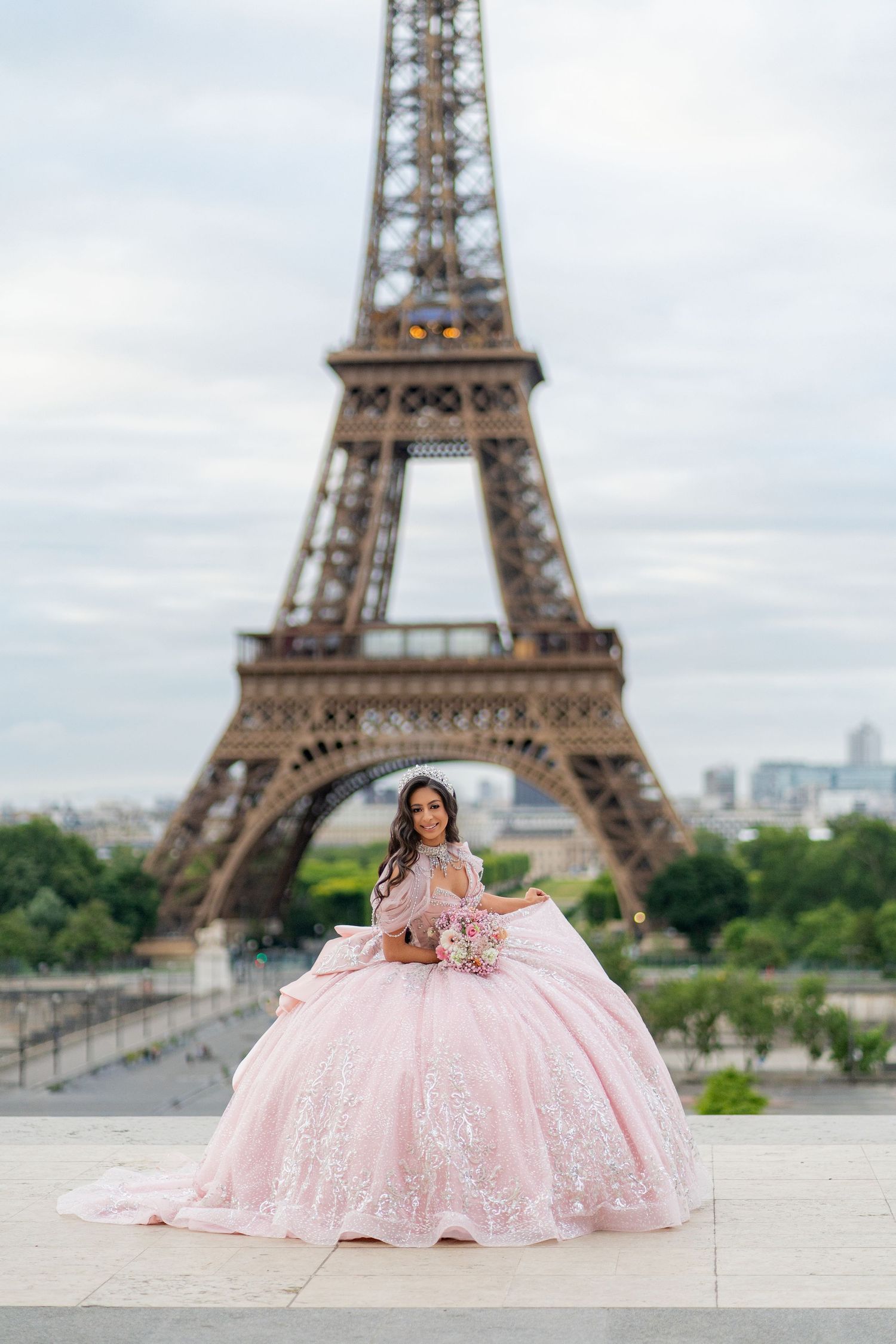 Young woman in pink lace ball gown poses with bouquet before iconic Eiffel Tower in Paris.