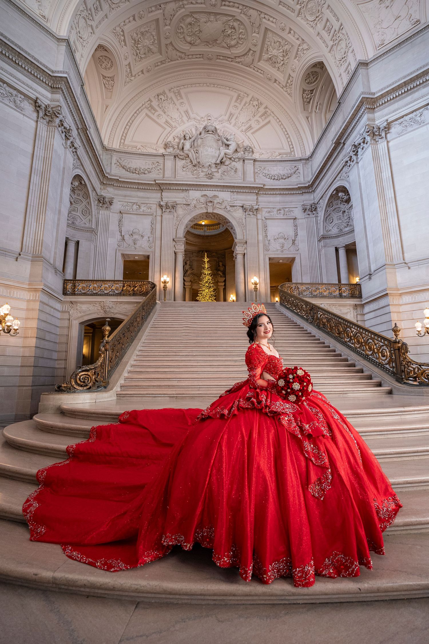 Young woman in stunning red ball gown with long train poses on grand marble staircase.