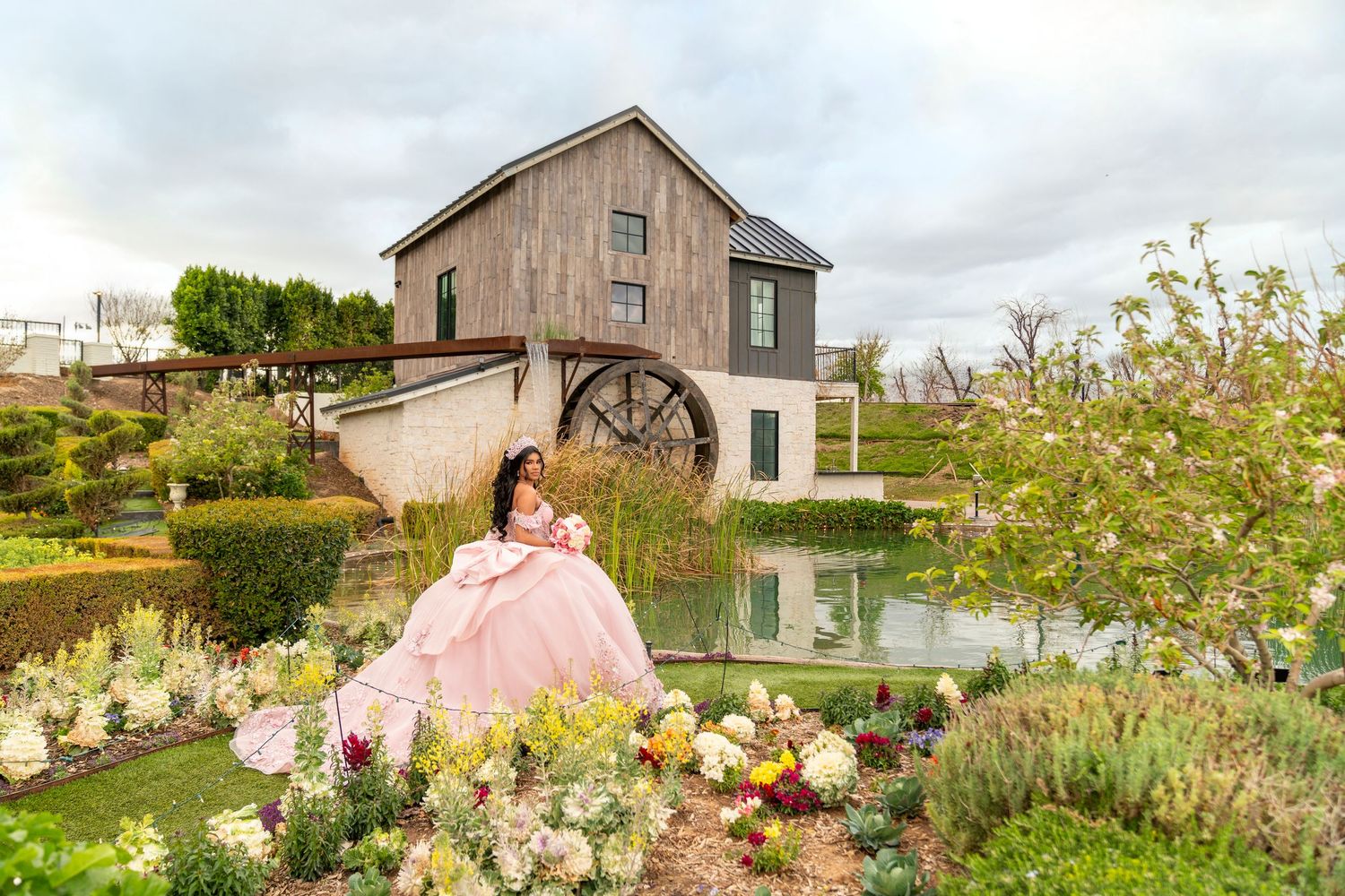 Woman in pink ball gown poses by a garden pond with a rustic mill backdrop and colorful flowers.
