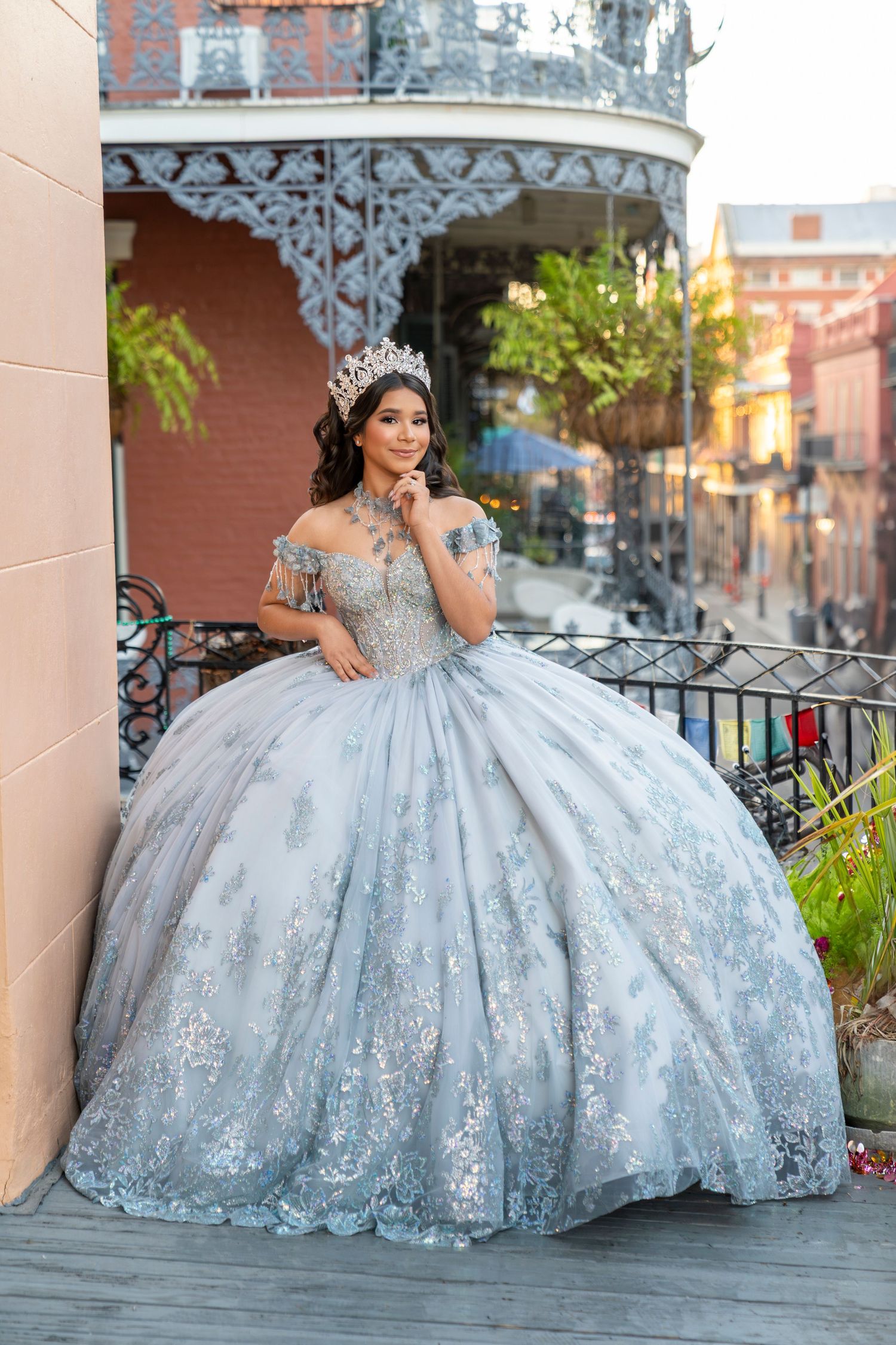 Young woman in silver quinceañera ball gown with floral lace and crown poses on balcony in historic district.