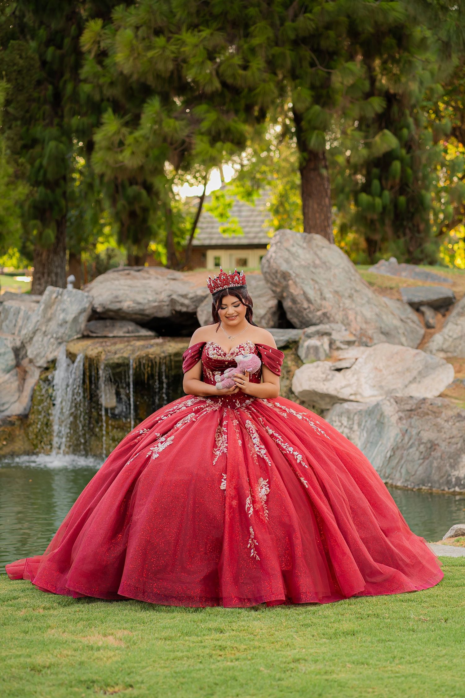Young woman in red ball gown and crown poses by waterfall for quinceañera celebration outdoors.