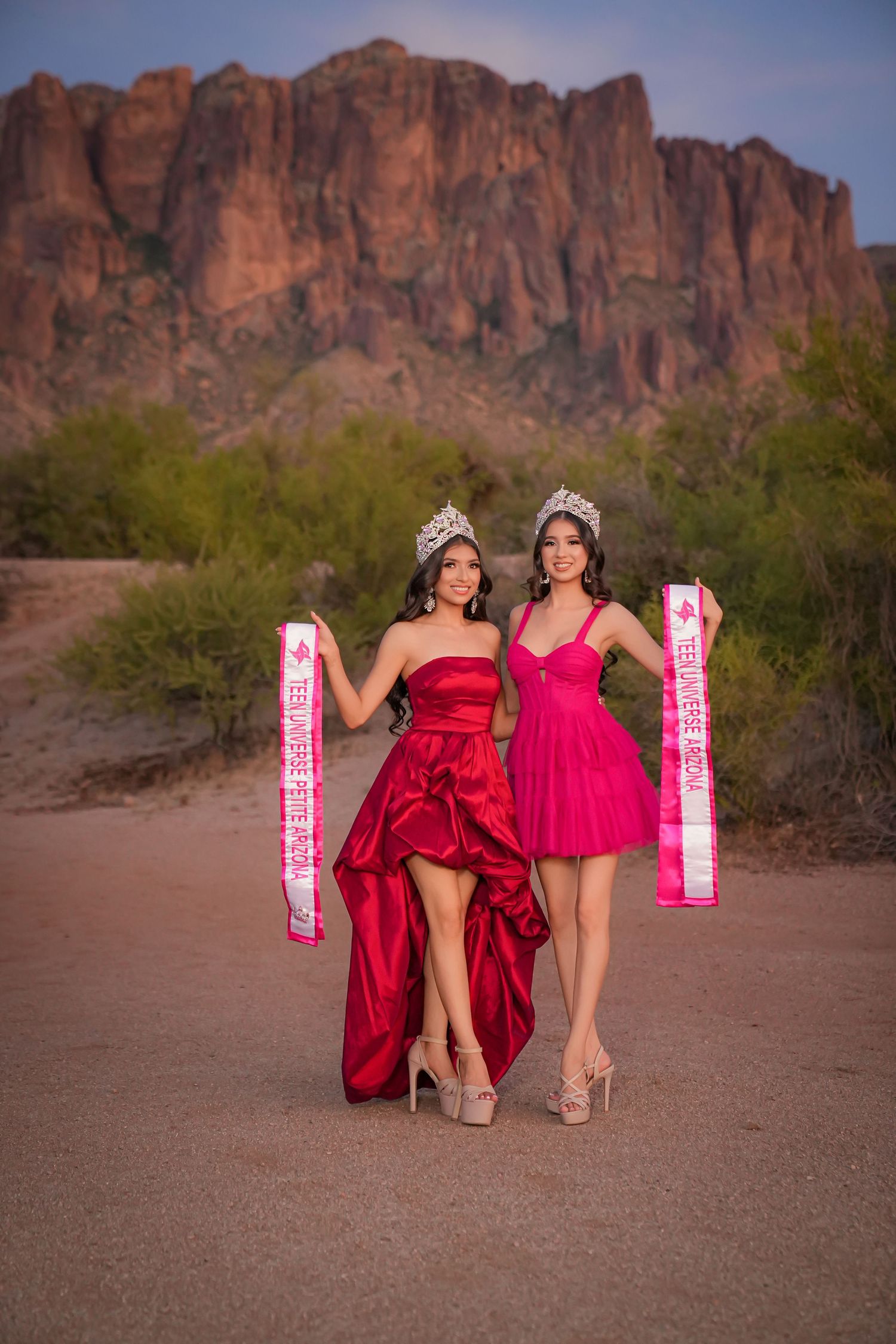 Two beauty queens in red and pink gowns with crowns and sashes pose before Superstition Mountains at sunset.