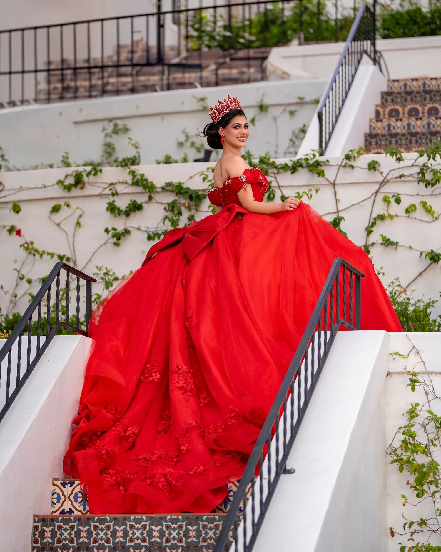 Young woman in a stunning red ball gown and crown poses gracefully on white stairs for her quinceañera.
