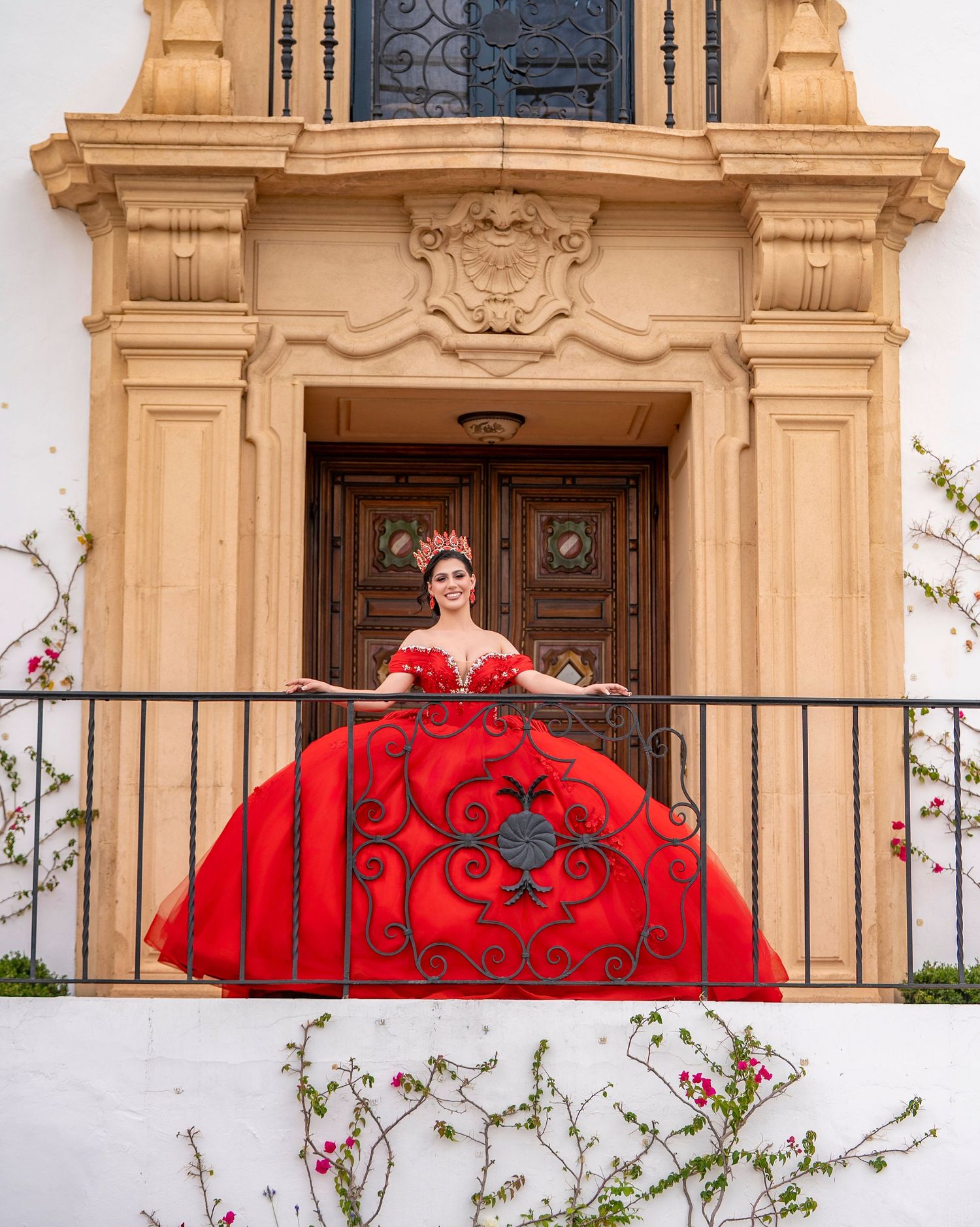 Young woman in a stunning red ball gown and crown poses on a balcony of an ornate building for her Quinceañera.