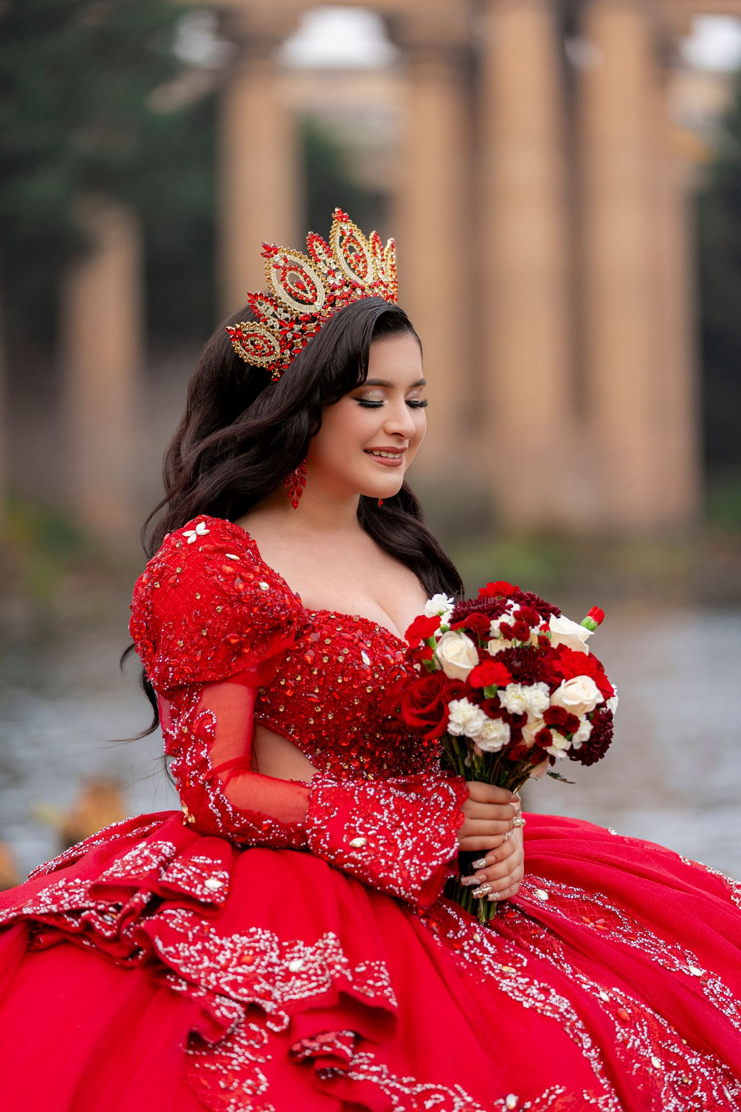 Young woman in red quinceañera gown with gold crown holds red and white bouquet, smiling outdoors.
