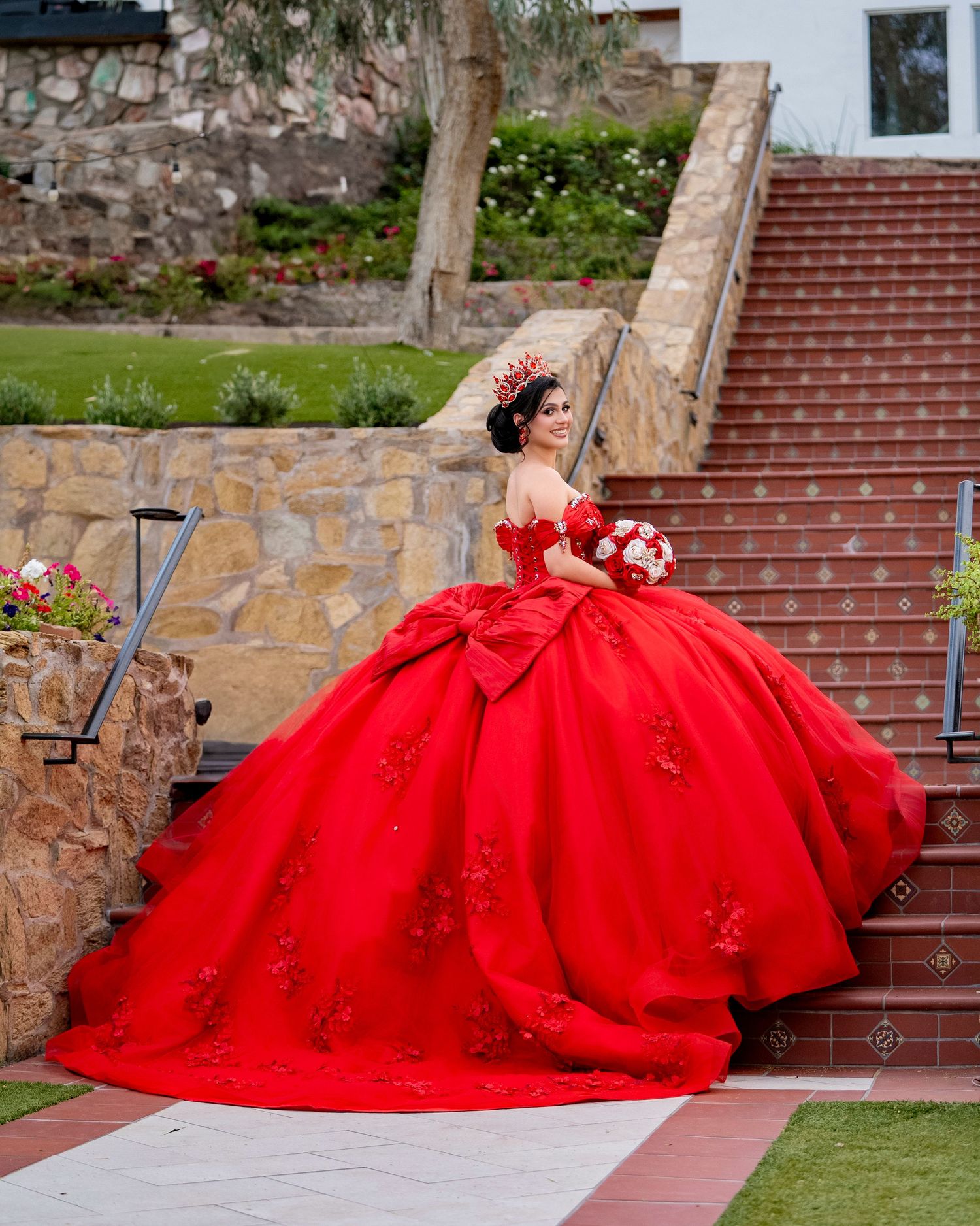 Beautiful quinceañera in a red off-shoulder ball gown with floral bouquet poses on stone staircase.