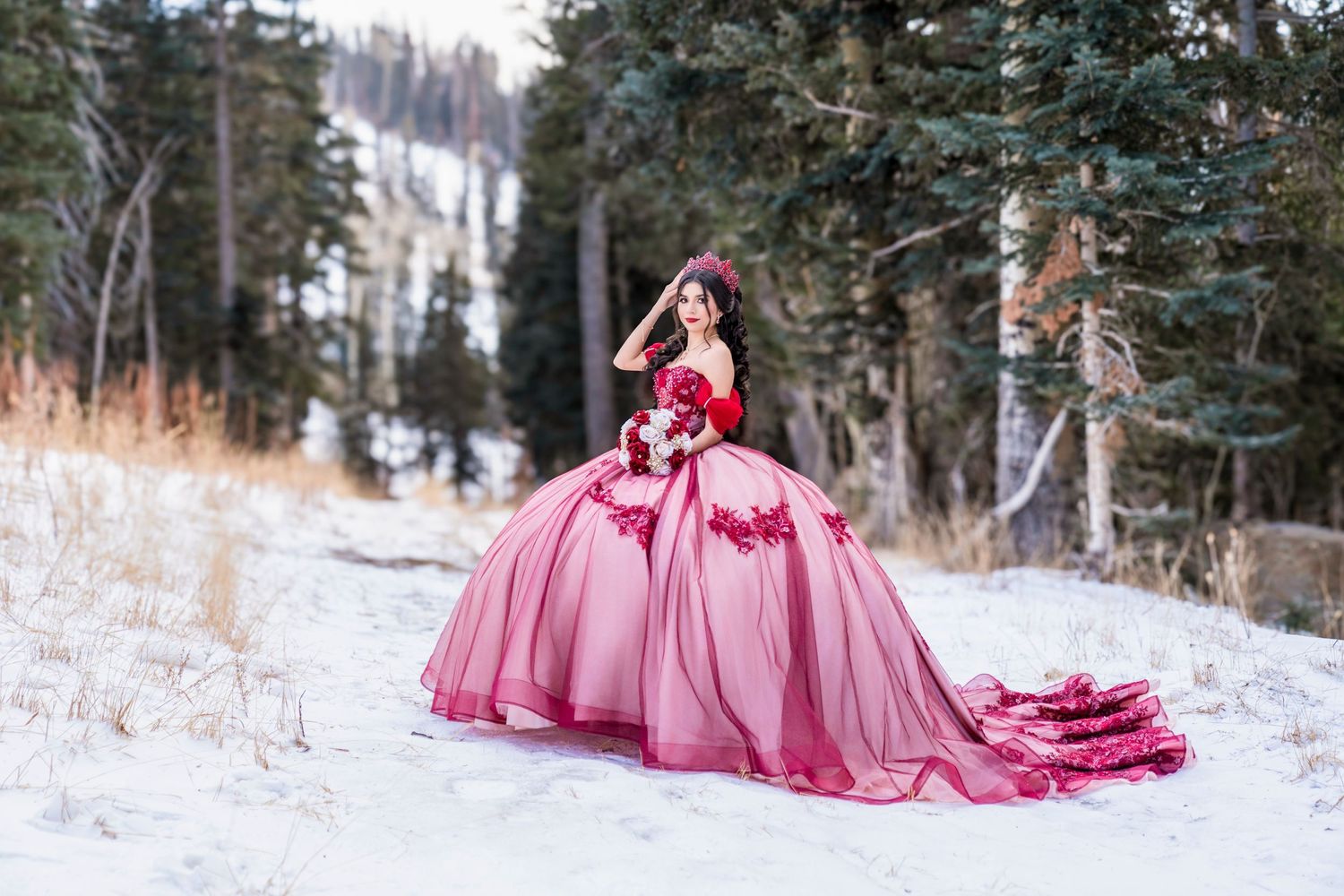 Woman in a dramatic pink and red ball gown with long train poses gracefully in a snowy pine forest.