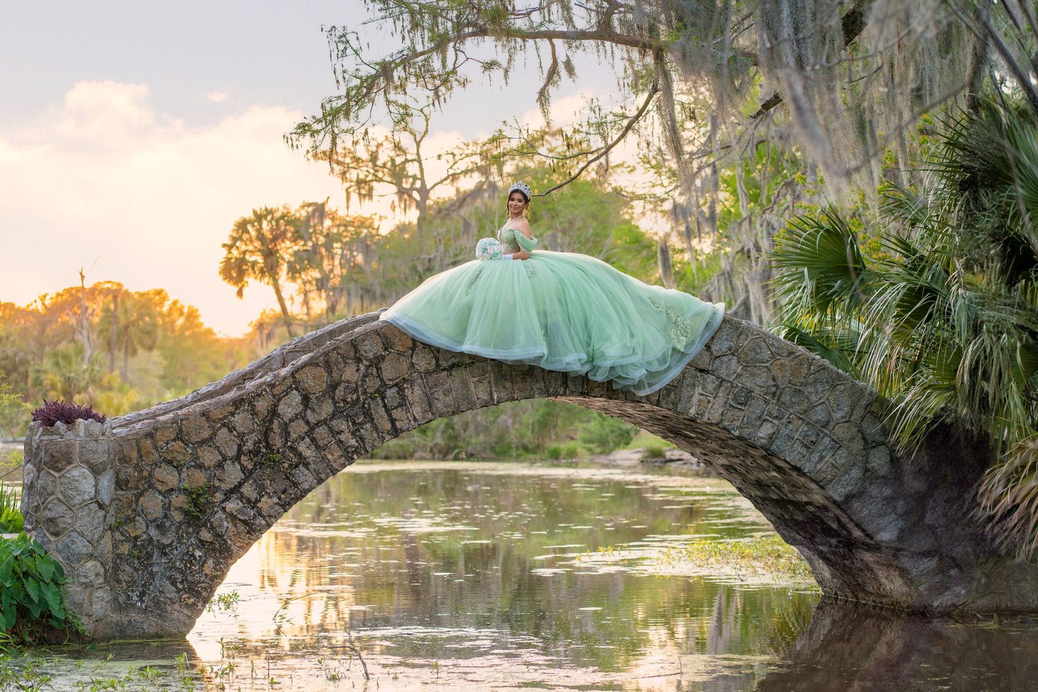 A young woman in a flowing mint green quinceañera gown poses gracefully on a stone arch bridge over a serene pond.