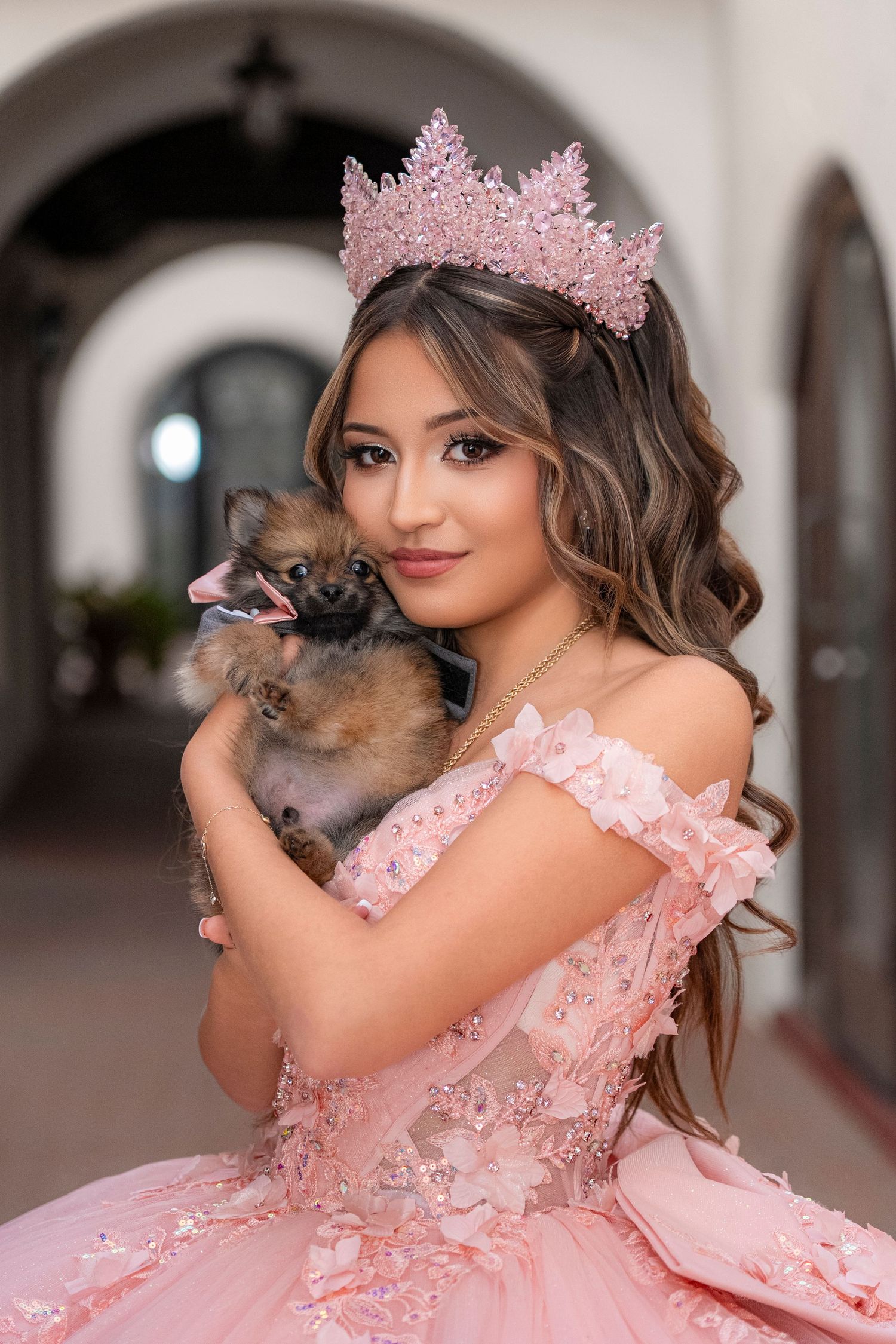 Young woman in pink quinceañera gown and crown holds a Pomeranian dog, posing elegantly in an arched hallway.