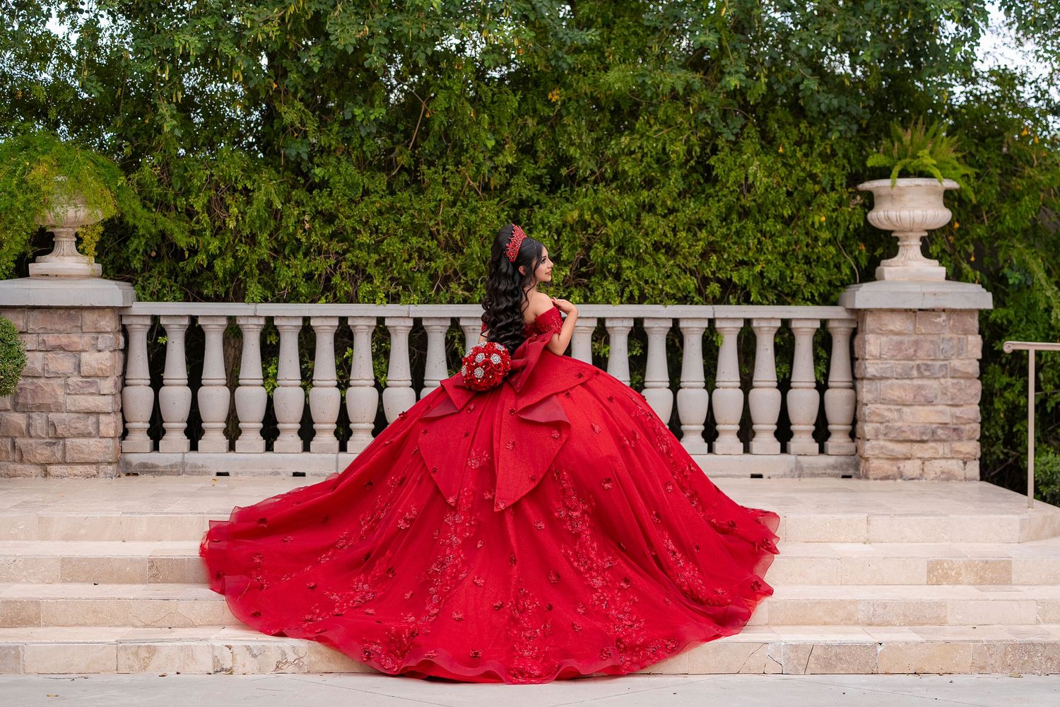 Elegant red ball gown with lace train spread on stone terrace steps, perfect for quinceañera or formal event.