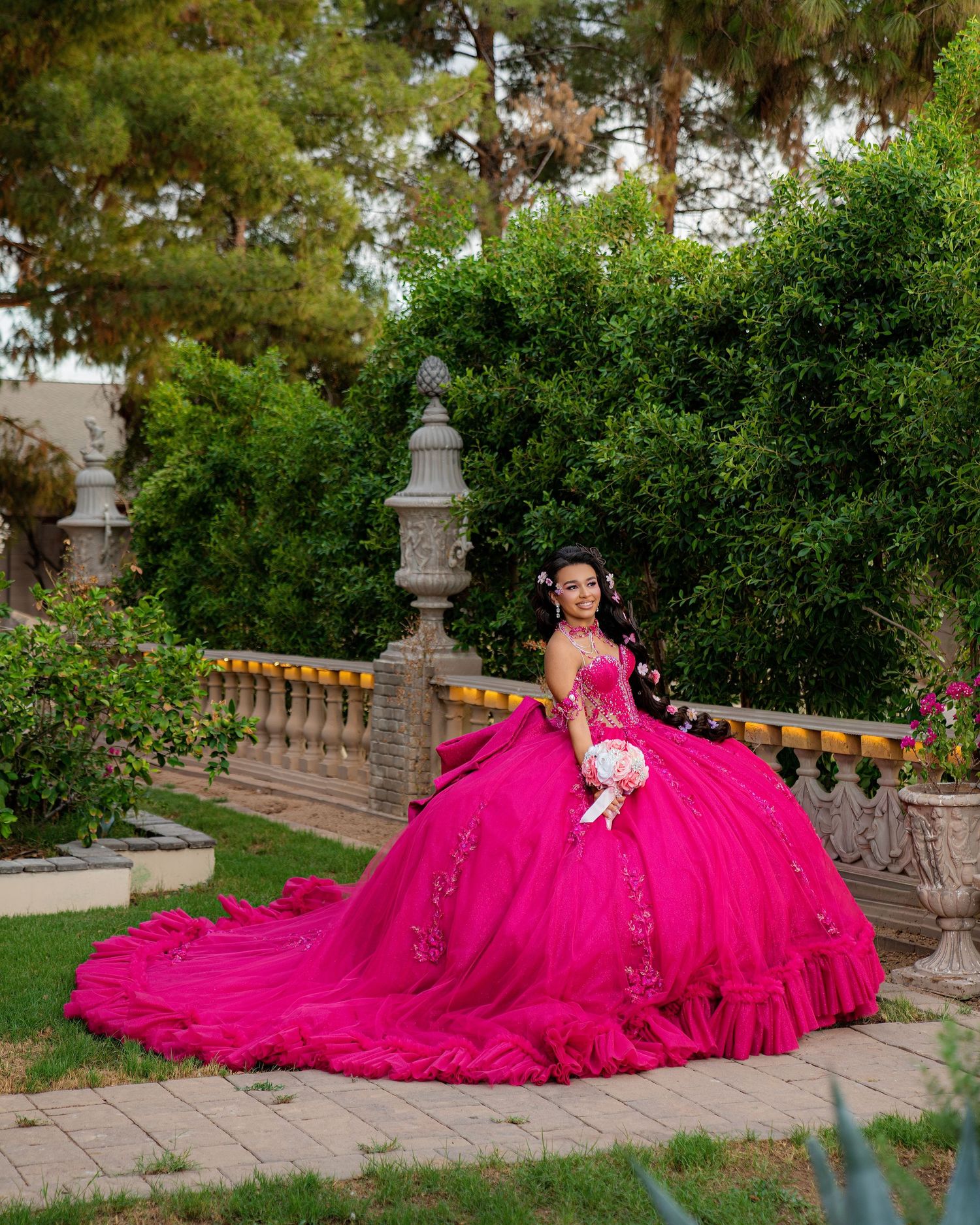 Young woman in a vibrant fuchsia quinceañera ball gown with floral details poses in a lush garden setting.