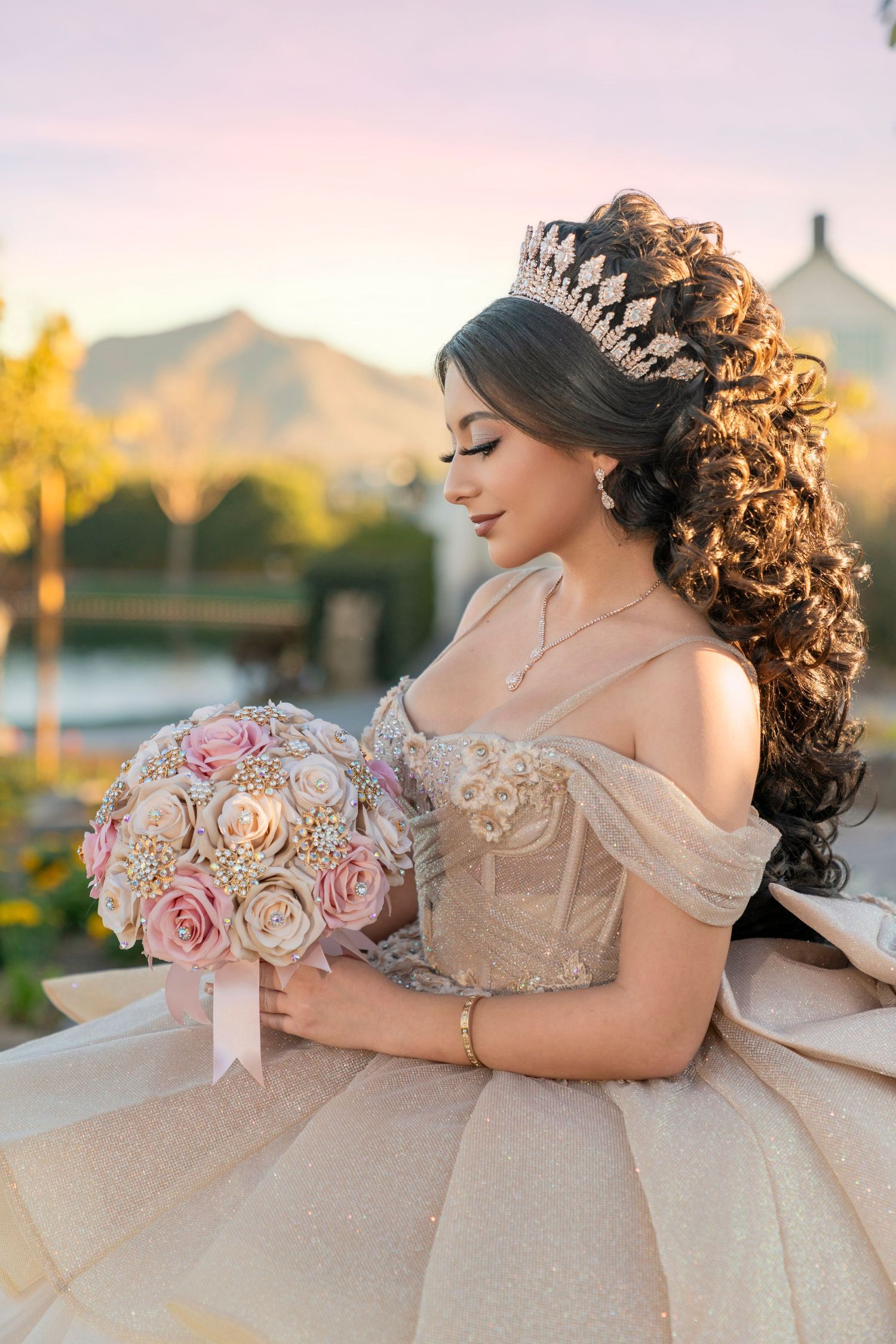 Young Latina in a champagne quinceañera gown with tiara holds a floral bouquet at golden hour outdoors.