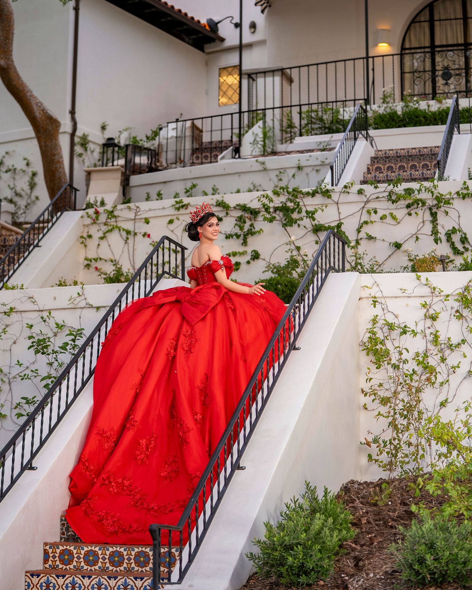 Young woman in dramatic red quinceañera gown drapes across white staircase of elegant estate.