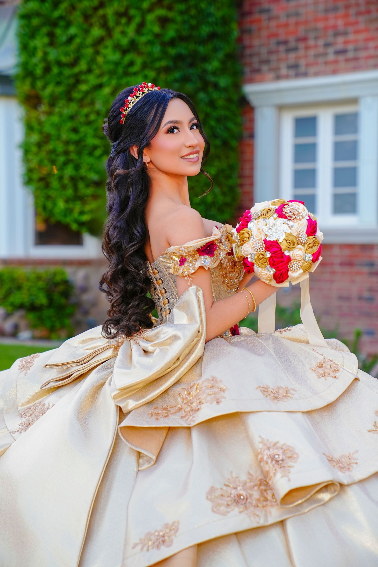 Smiling young woman in a gold quinceañera gown holding a red and gold floral bouquet outdoors.