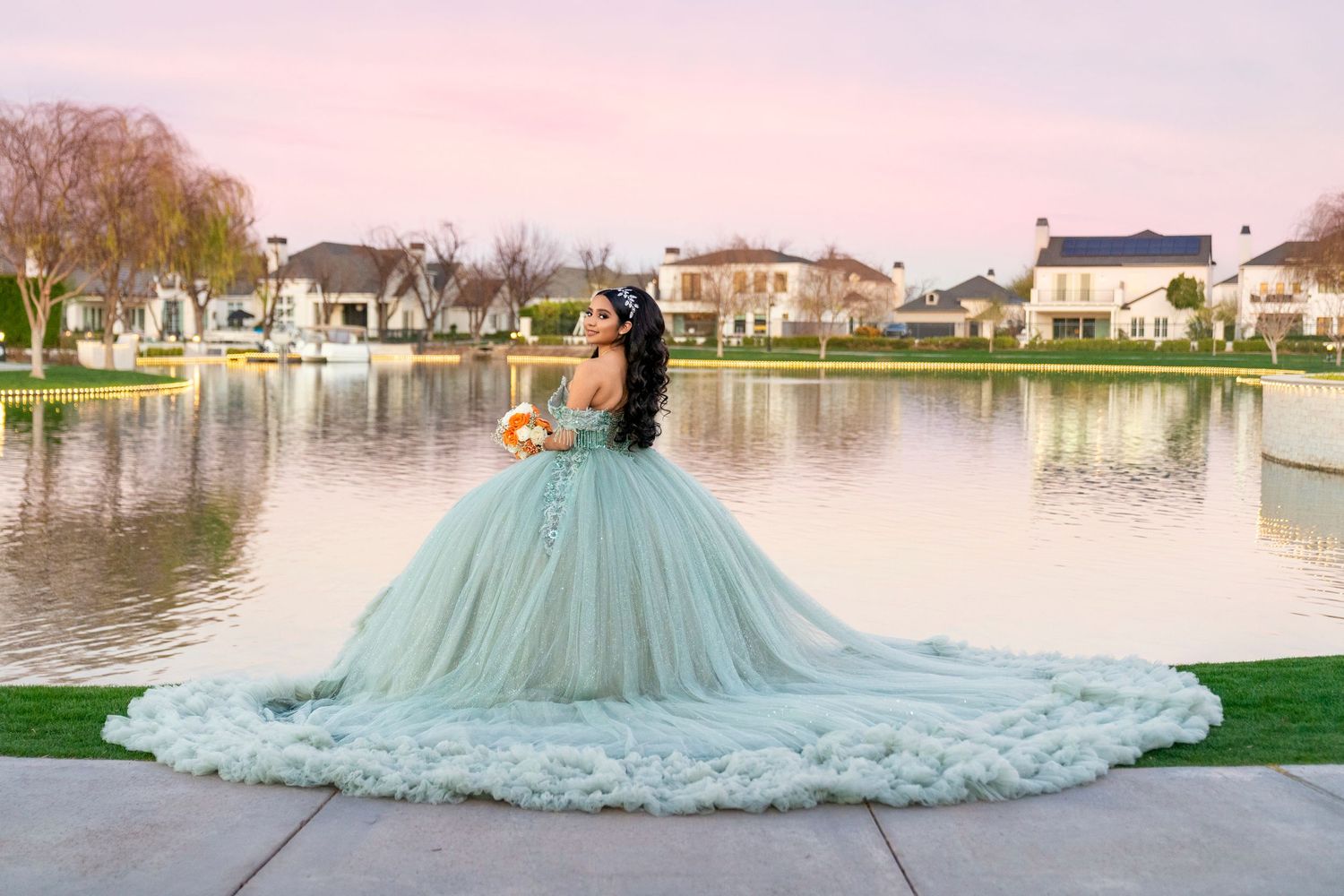 A woman in a stunning mint green ball gown poses by a serene pond at sunset in an elegant suburban setting.