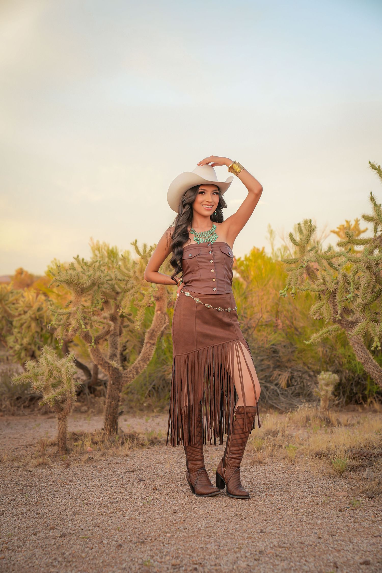 A woman in a brown fringe dress, cowboy hat, and boots poses in a desert landscape with cacti at sunset.