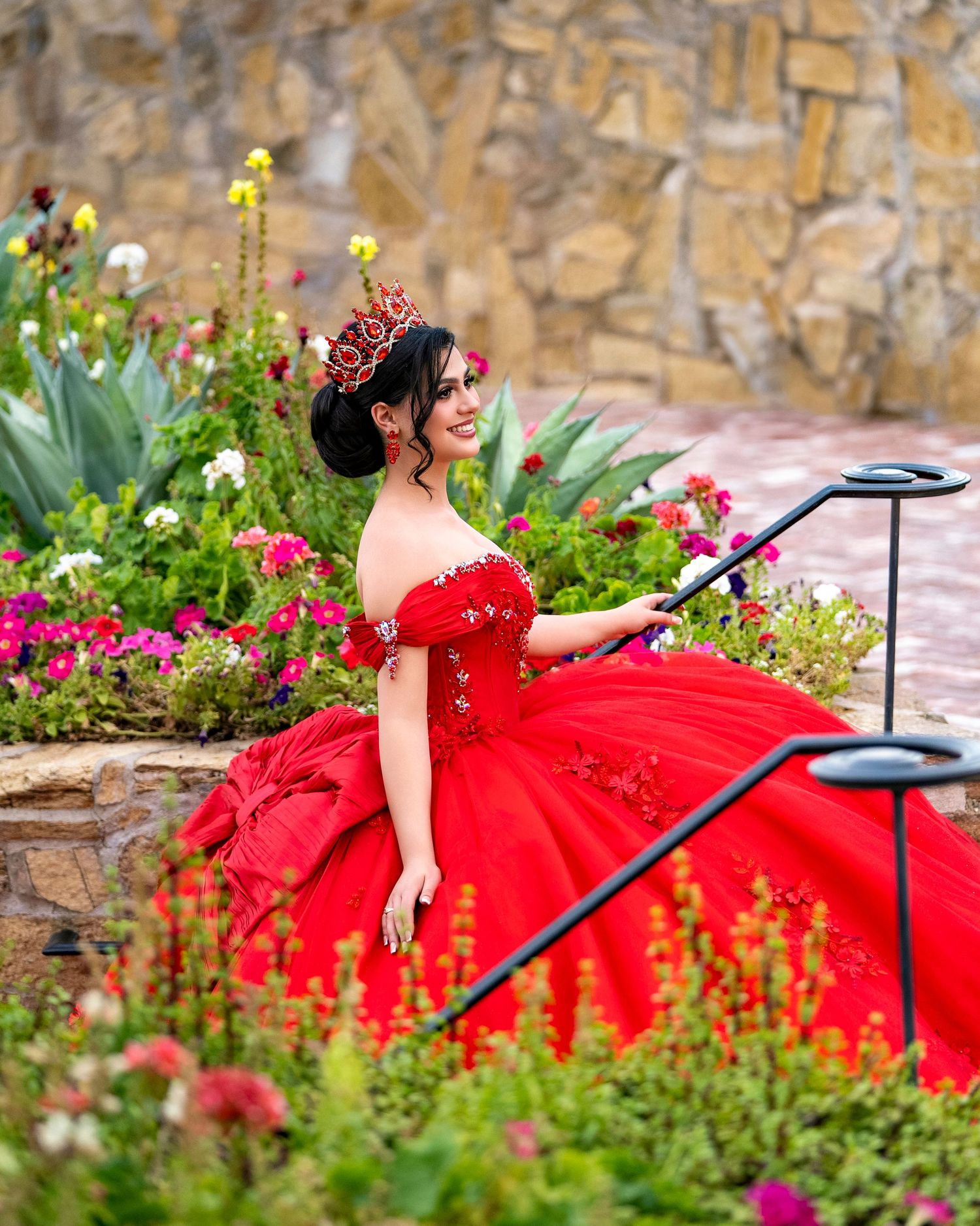 Young woman in a red ball gown and crown poses gracefully in a flower garden for her quinceañera.