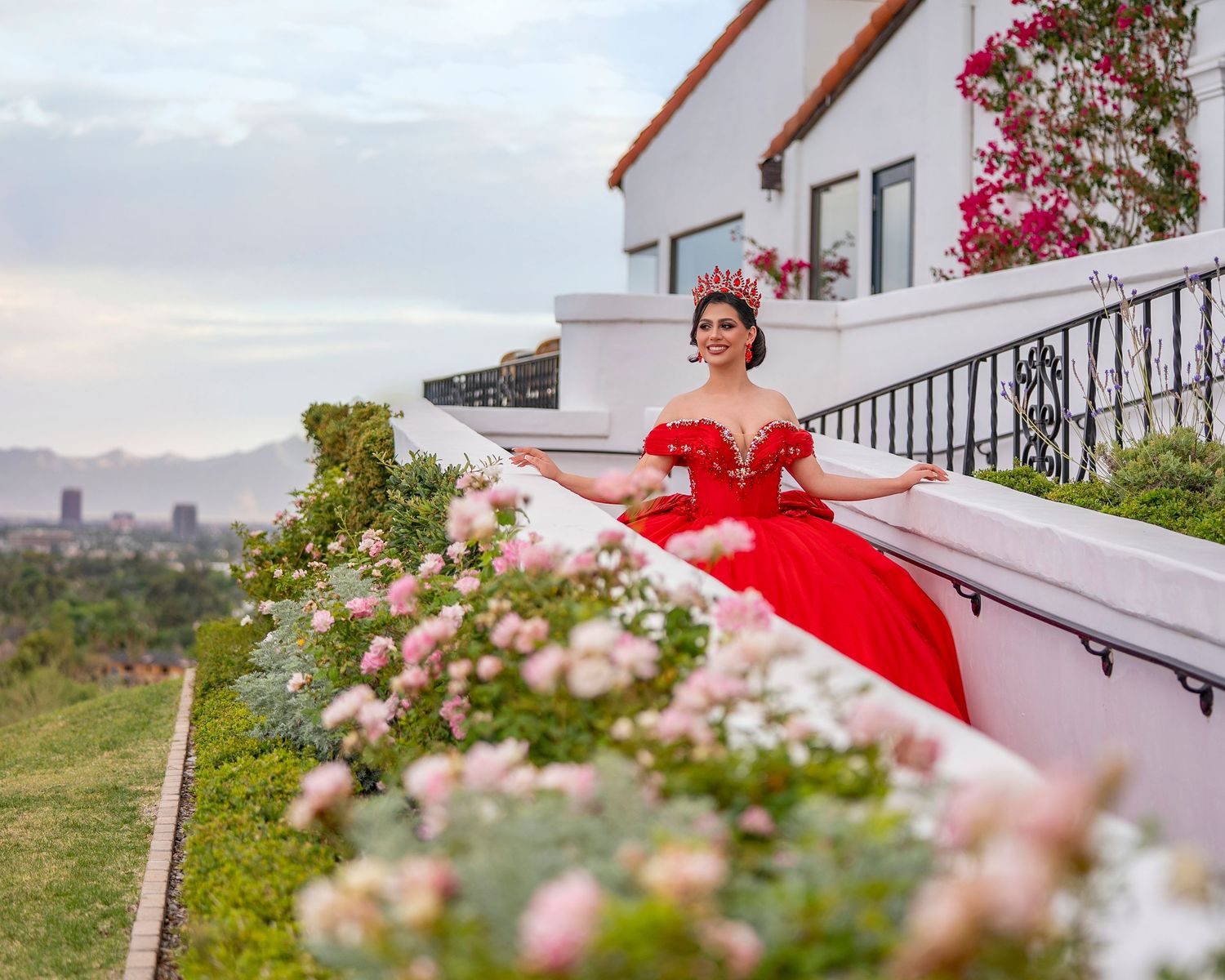 Young woman in red quinceañera gown reclines among pink roses with city skyline in background.