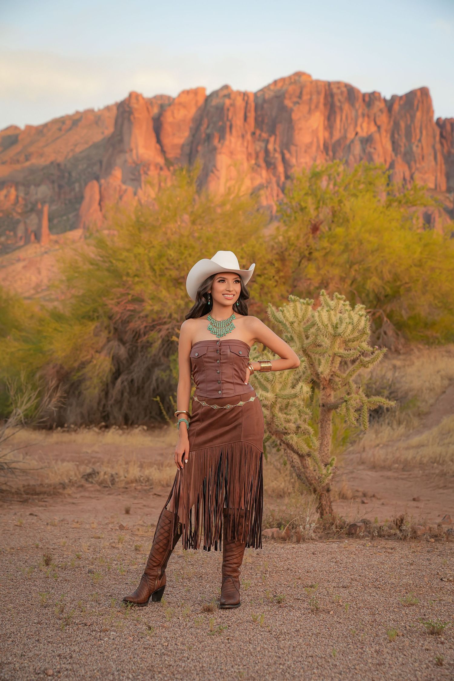 Woman in cowboy hat and brown fringe dress poses before red rock mountains and cacti at golden hour.