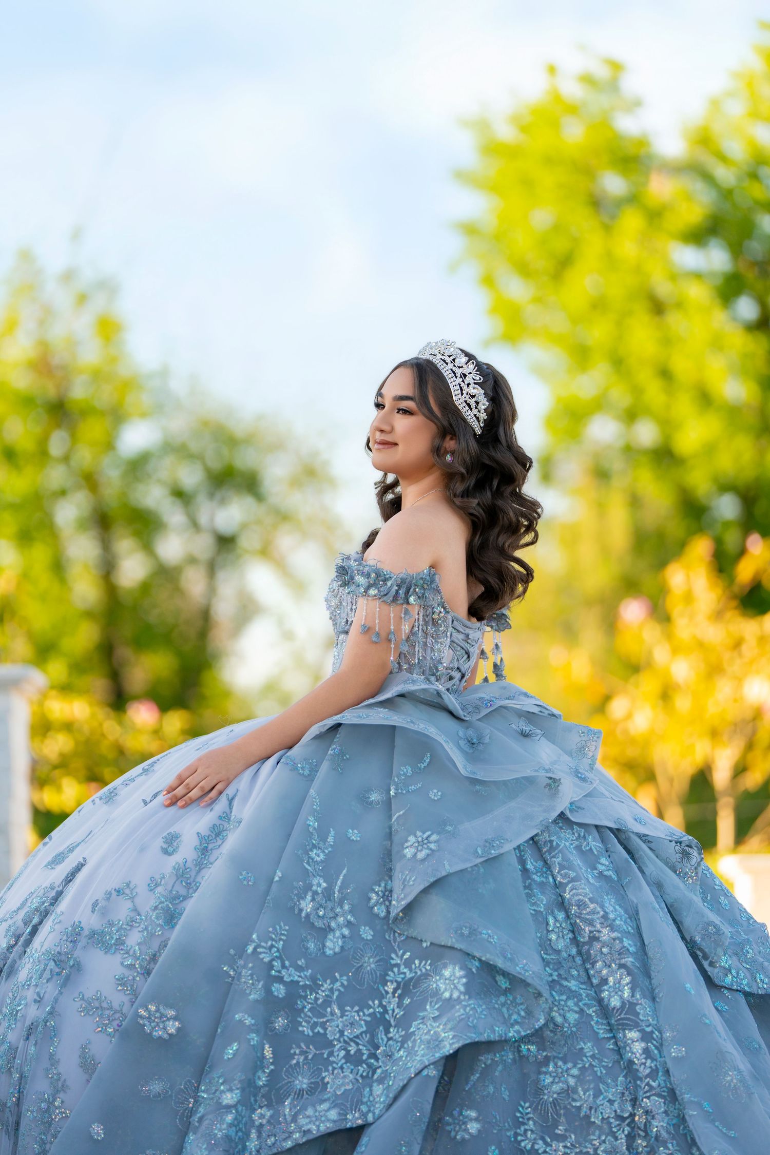 Young woman in a blue floral ball gown and tiara smiling outdoors at her quinceañera celebration.