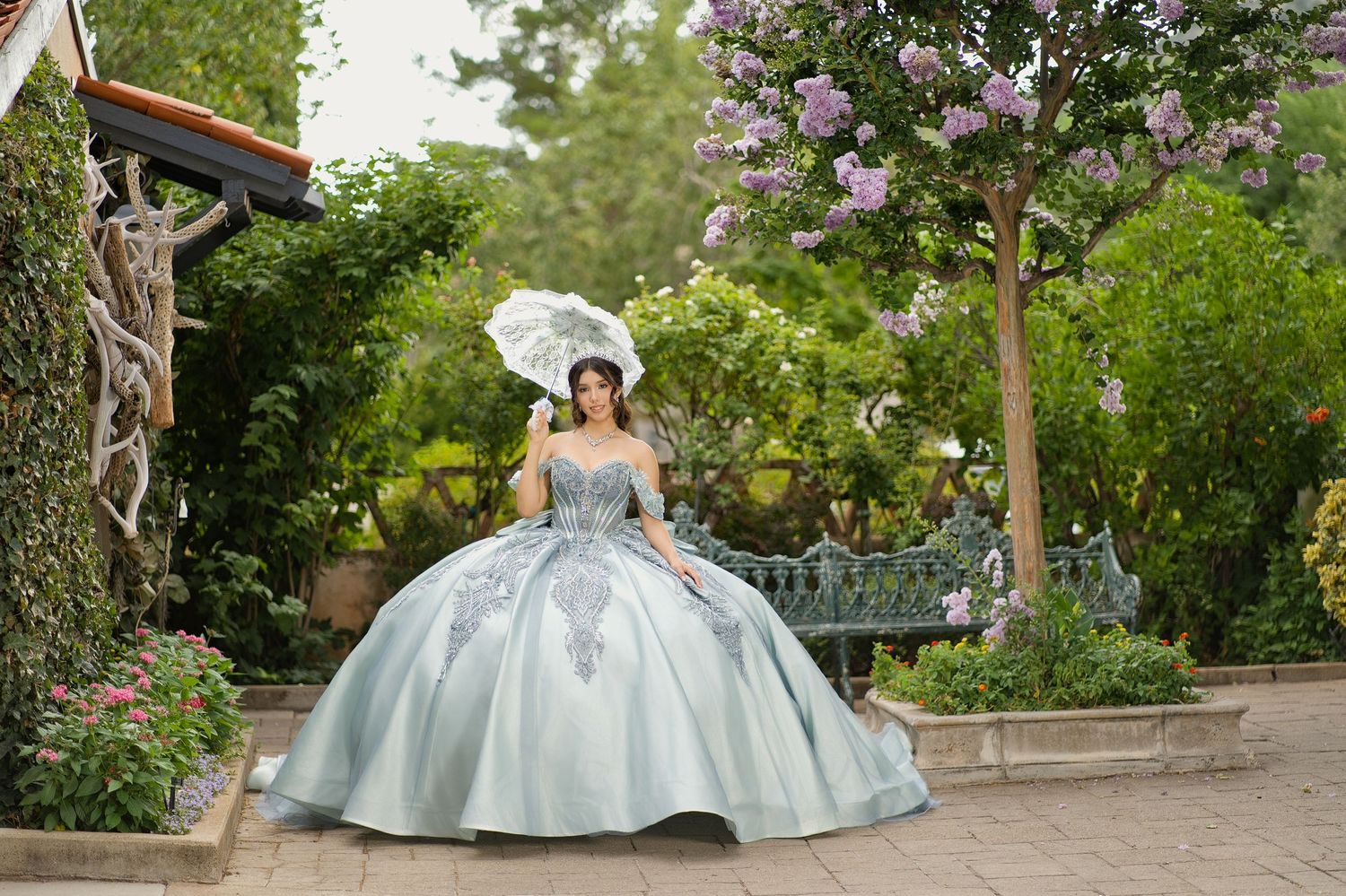 Young woman in a light blue ballgown quinceañera dress holding a white parasol in a lush garden setting.