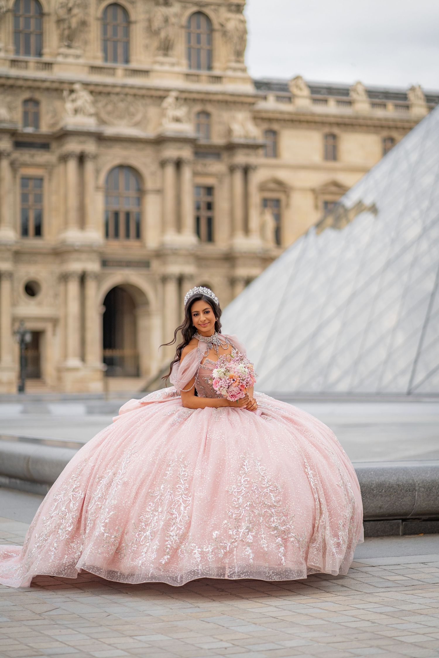 Young woman in pink quinceañera gown poses with bouquet near the Louvre Pyramid in Paris.