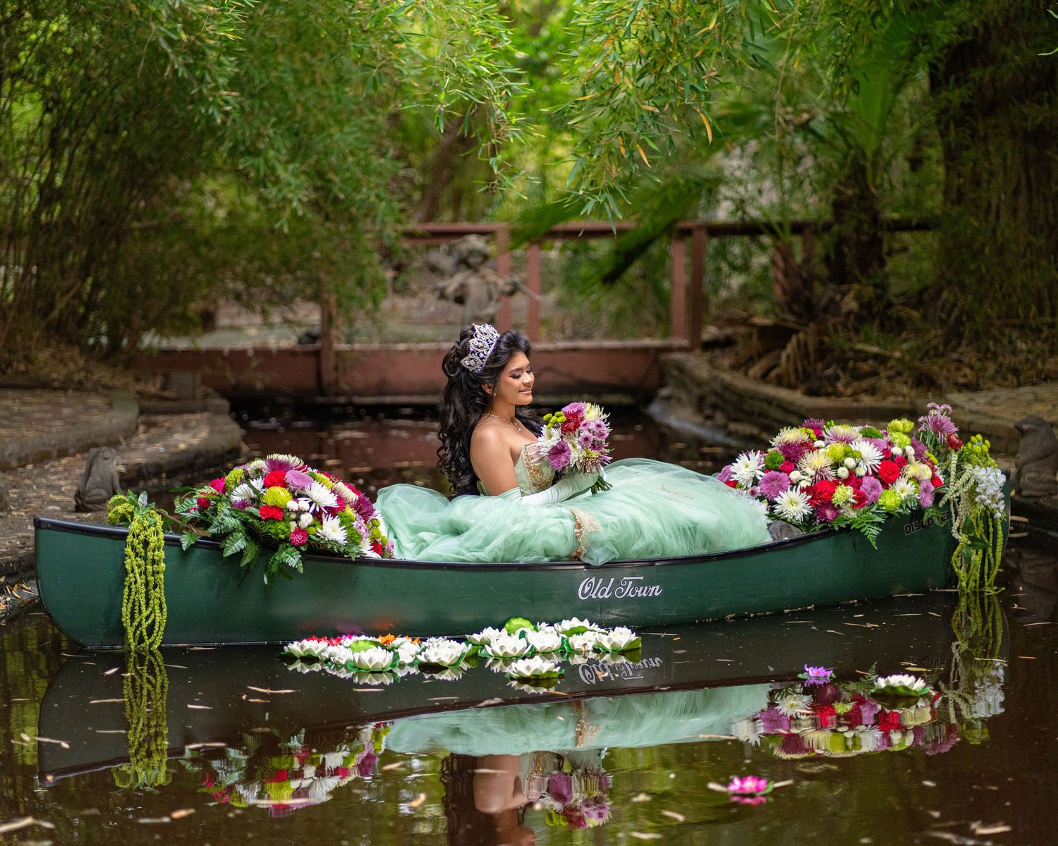 Young woman in mint green ball gown holds colorful bouquet while sitting in floral-decorated green canoe.