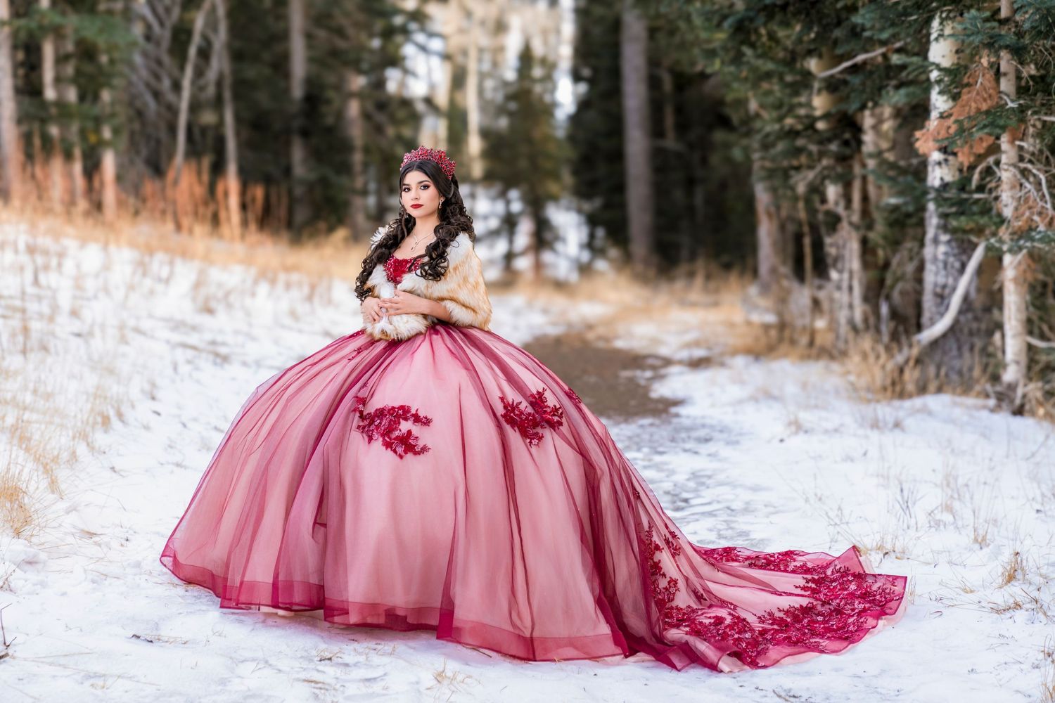 Young woman in a stunning burgundy ball gown quinceañera dress posing in a snowy winter forest setting.
