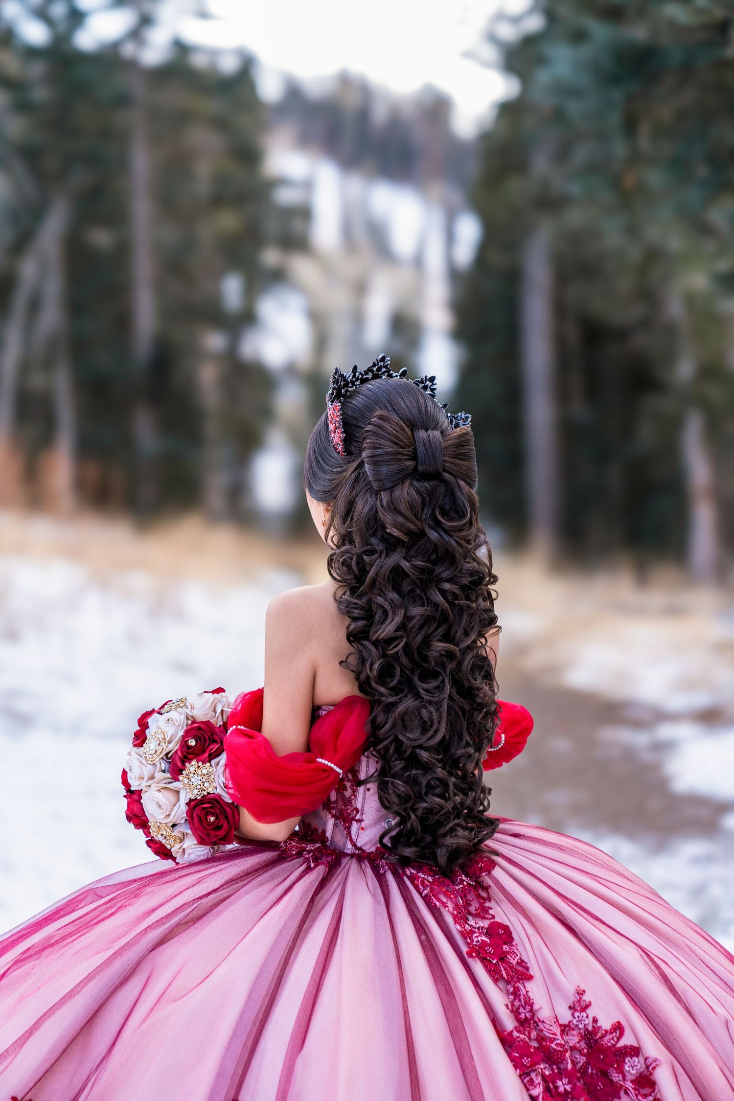 Quinceañera in pink and red ball gown holds bouquet in snowy forest.