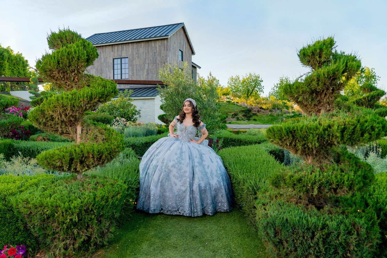 A woman in an elegant blue ball gown poses in a manicured garden with sculpted hedges and a rustic house backdrop.