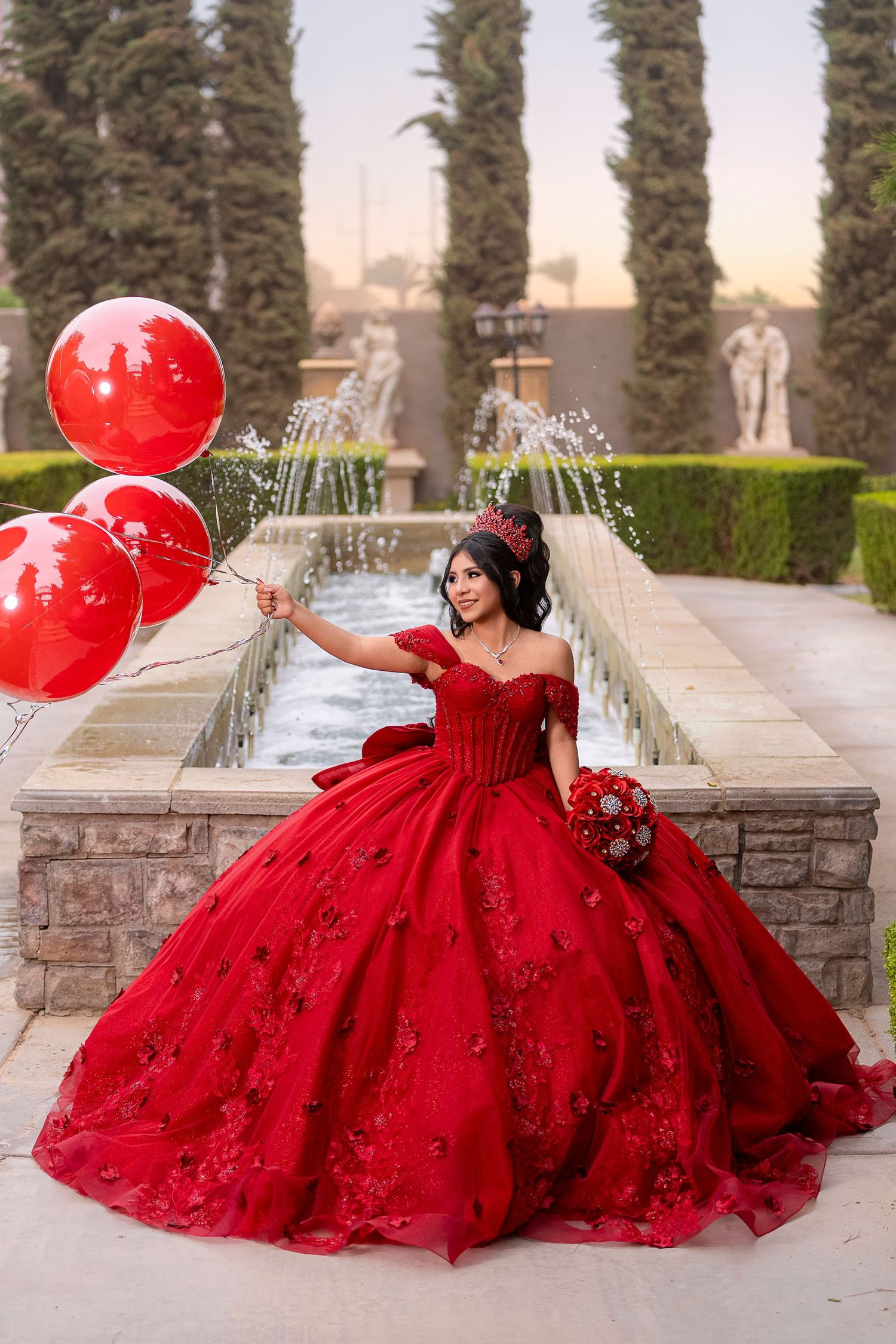Young woman in red ball gown quinceañera dress holds red balloons and bouquet in elegant garden setting.