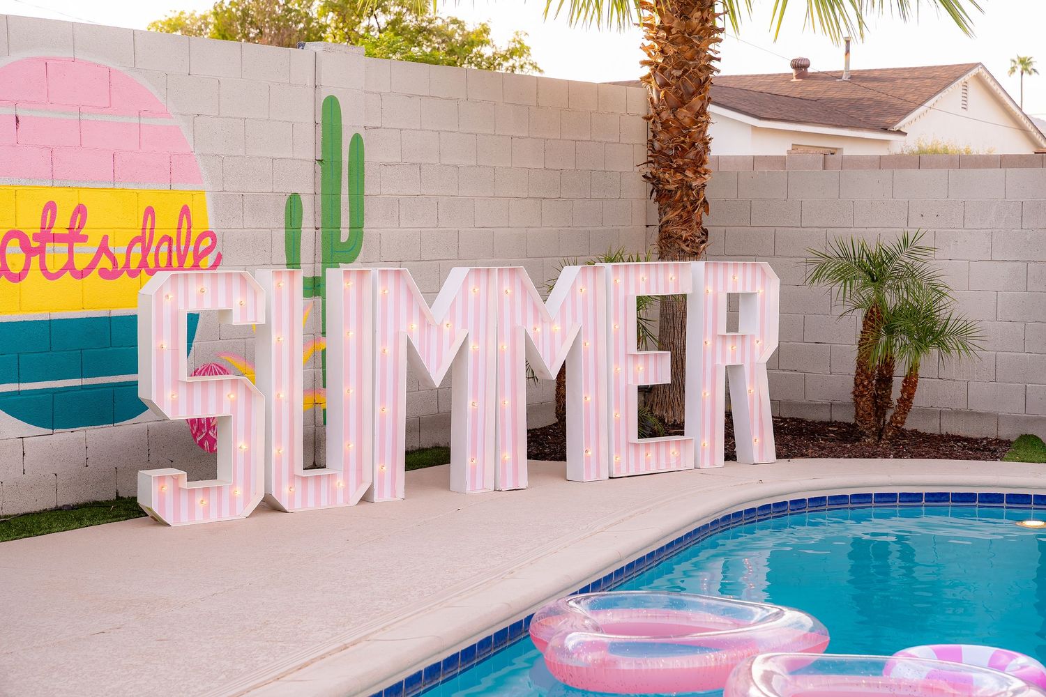 Pink SUMMER marquee letters by a Scottsdale pool with a cactus mural and colorful floaties on a sunny day.