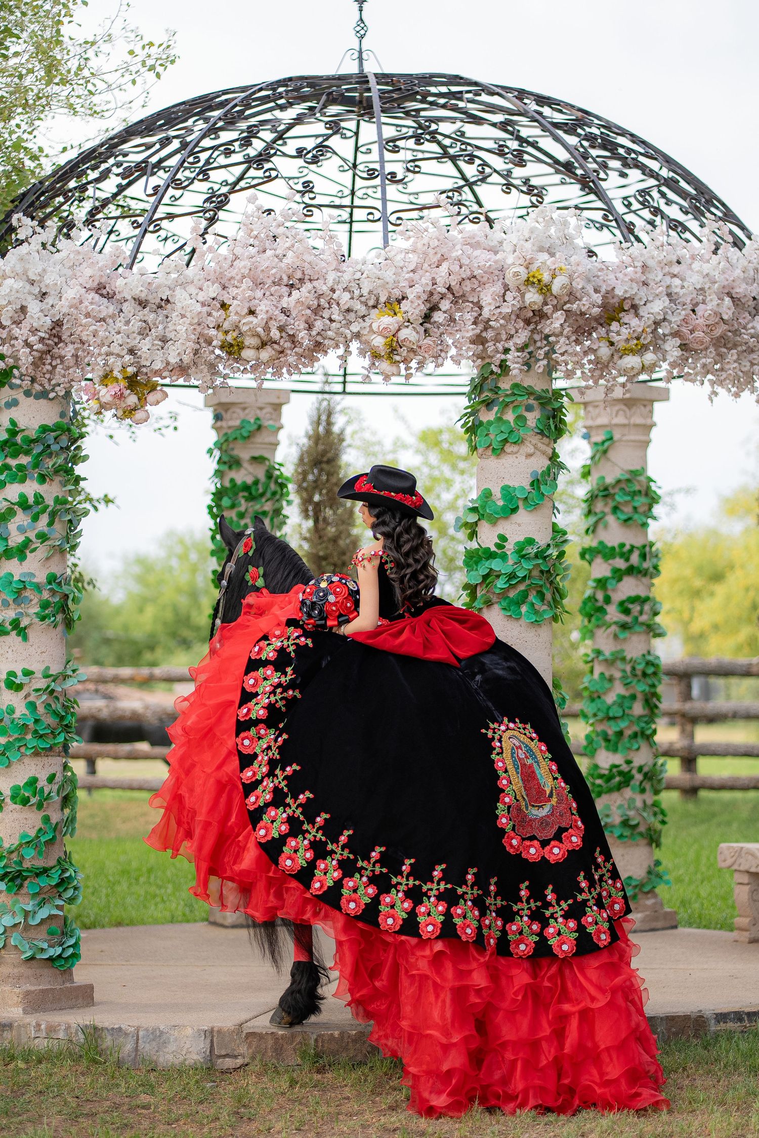 Woman in red and black Mexican dress with floral embroidery poses under a floral gazebo with ivy-wrapped columns.