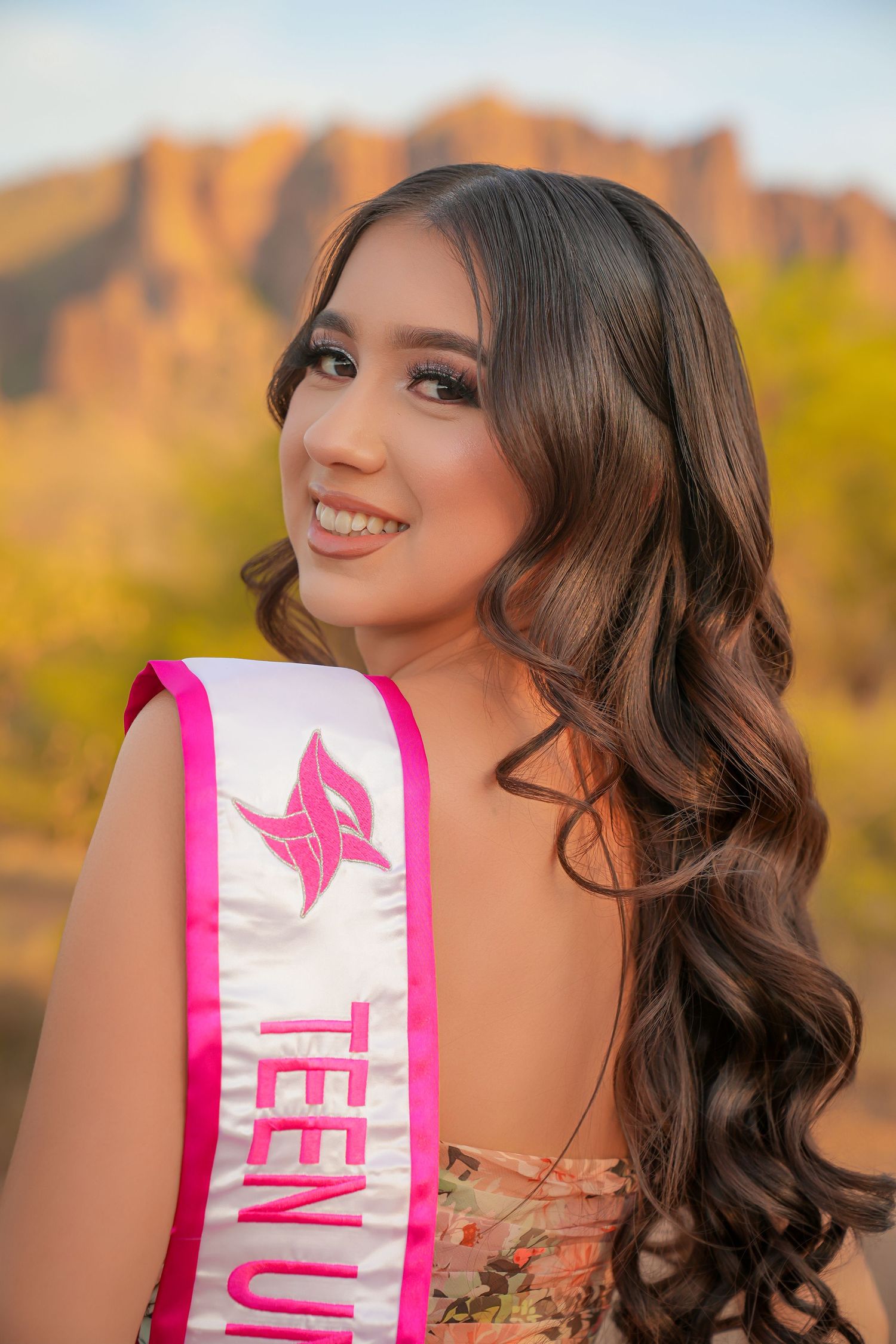 Teen beauty queen wearing a pink sash smiles outdoors with scenic desert rock formations in the golden hour background.