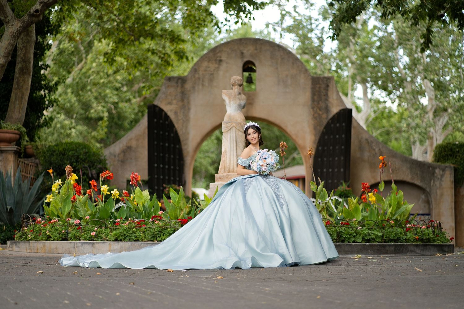 Young woman in a light blue ball gown quinceañera dress poses in a garden with colorful flowers and stone archway.