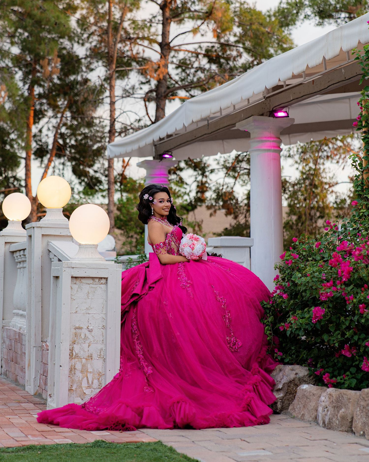 Young woman in a voluminous hot pink quinceañera ball gown poses gracefully by a white gazebo surrounded by flowers.