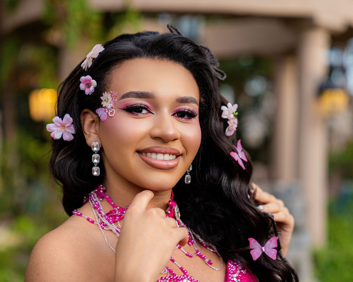 Young woman in pink beaded dress with floral hair accessories and butterfly clips, wearing pink eye makeup and drop earrings.