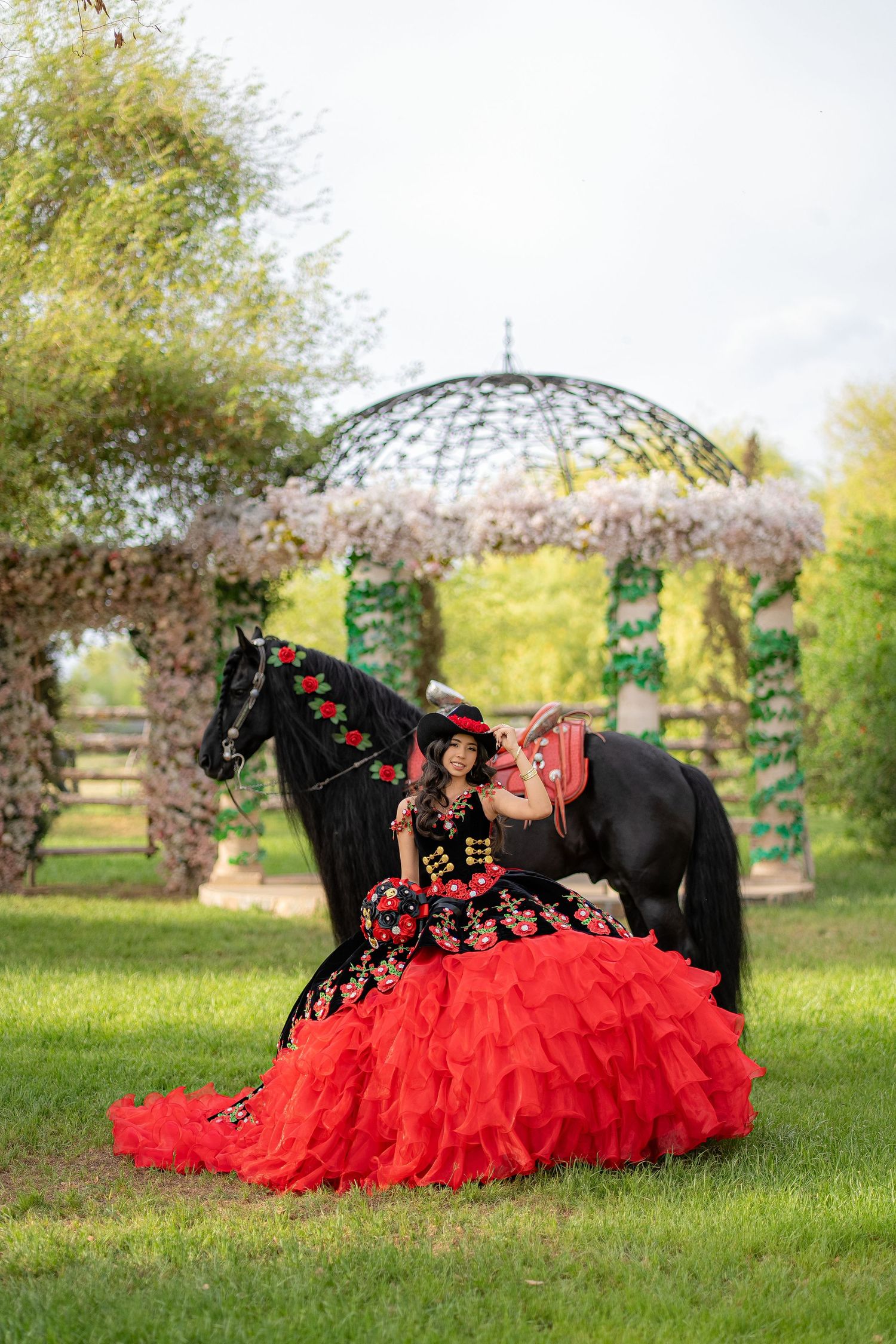 A woman in a red and black floral quinceañera gown poses with a black horse near a floral gazebo.