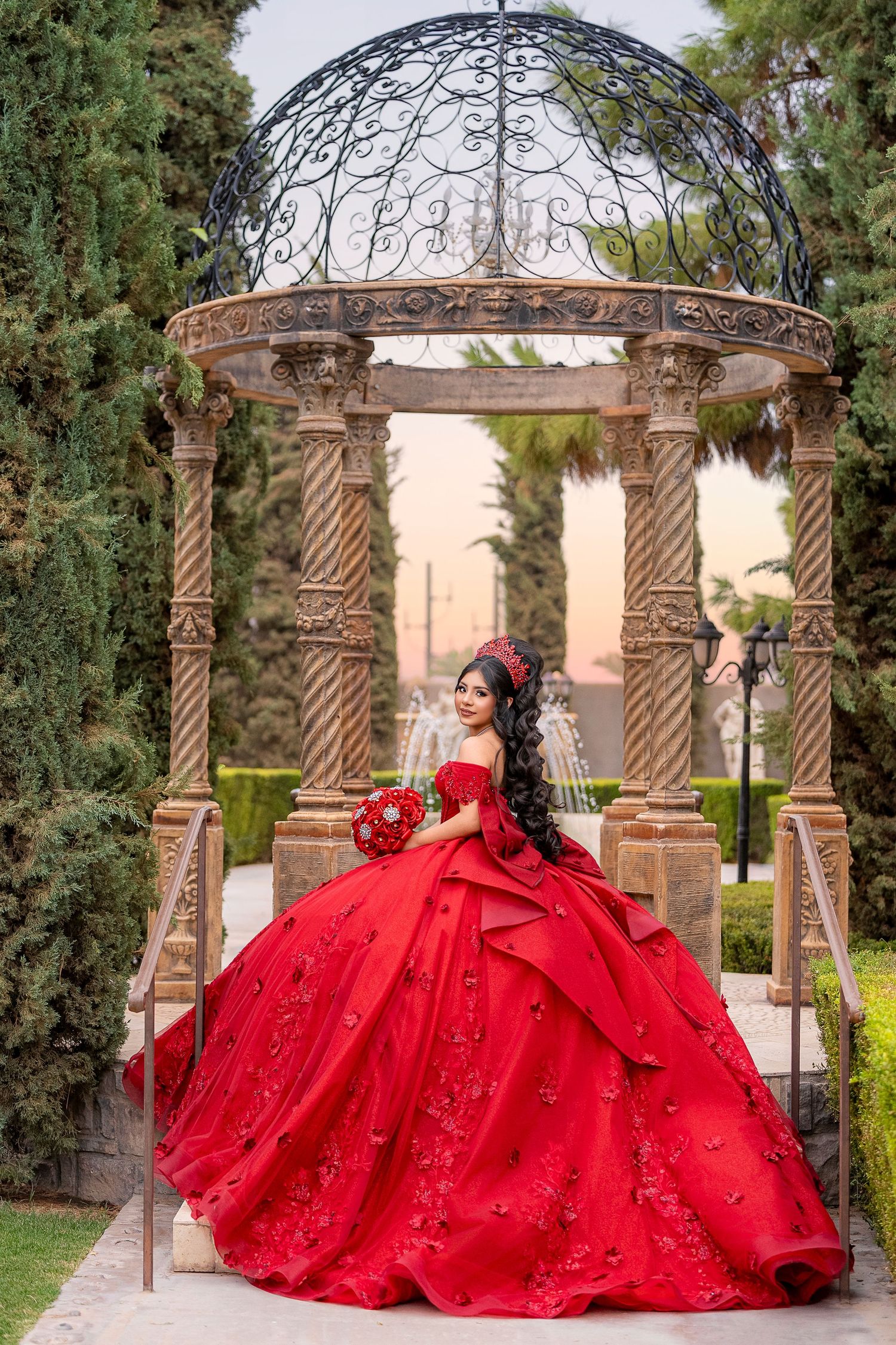 Young woman in a stunning red ball gown quinceañera dress poses under an ornate gazebo in a lush garden.