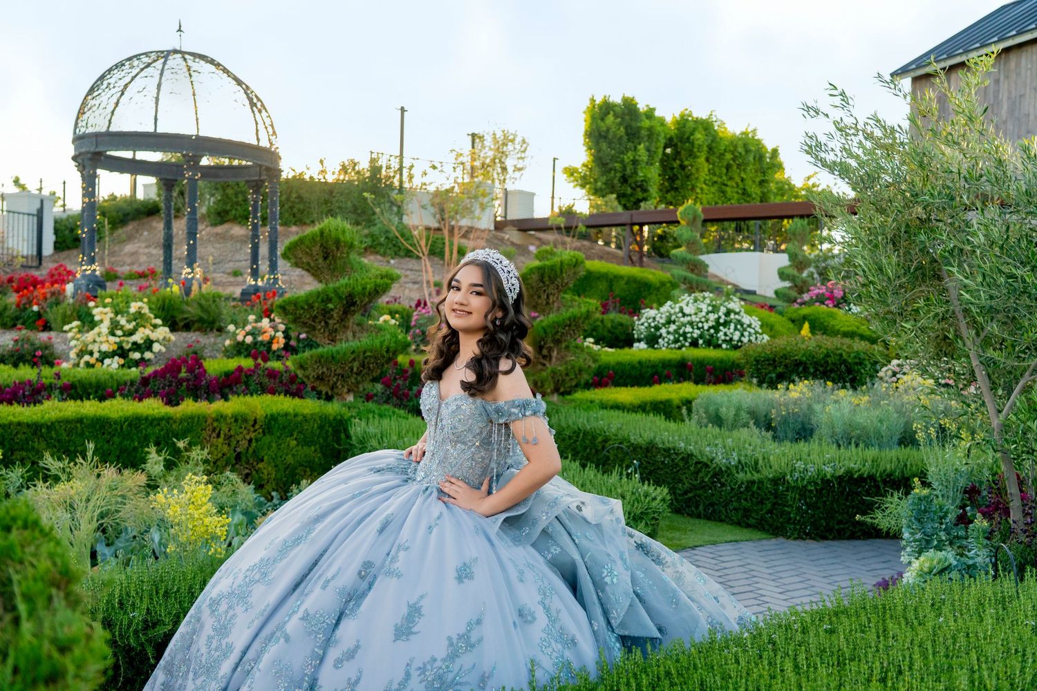 Young woman in a light blue quinceañera ball gown poses in a lush formal garden with a gazebo backdrop.