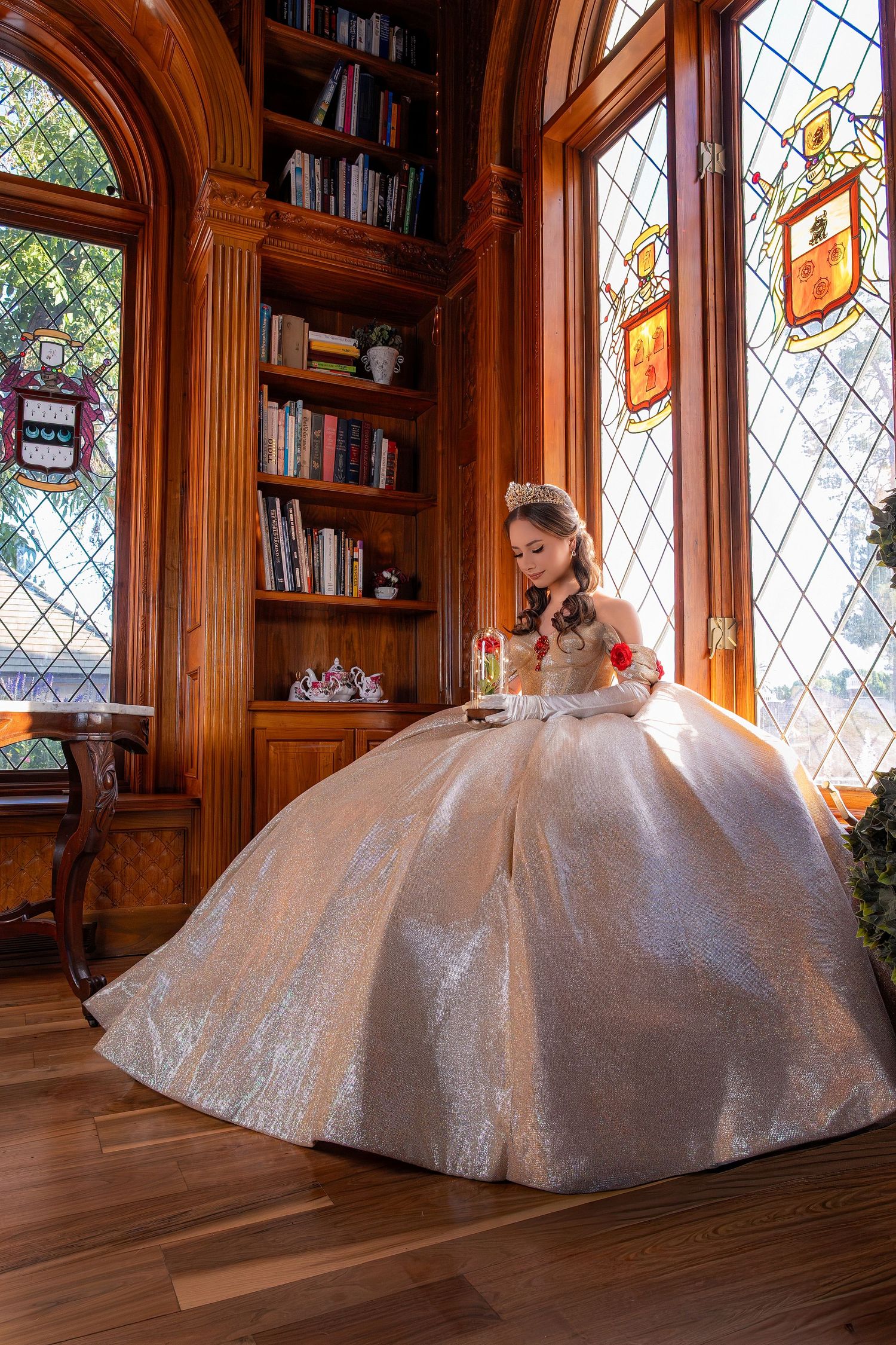 Young woman in a shimmering white ball gown with tiara poses in an elegant wood-paneled library with stained glass windows.