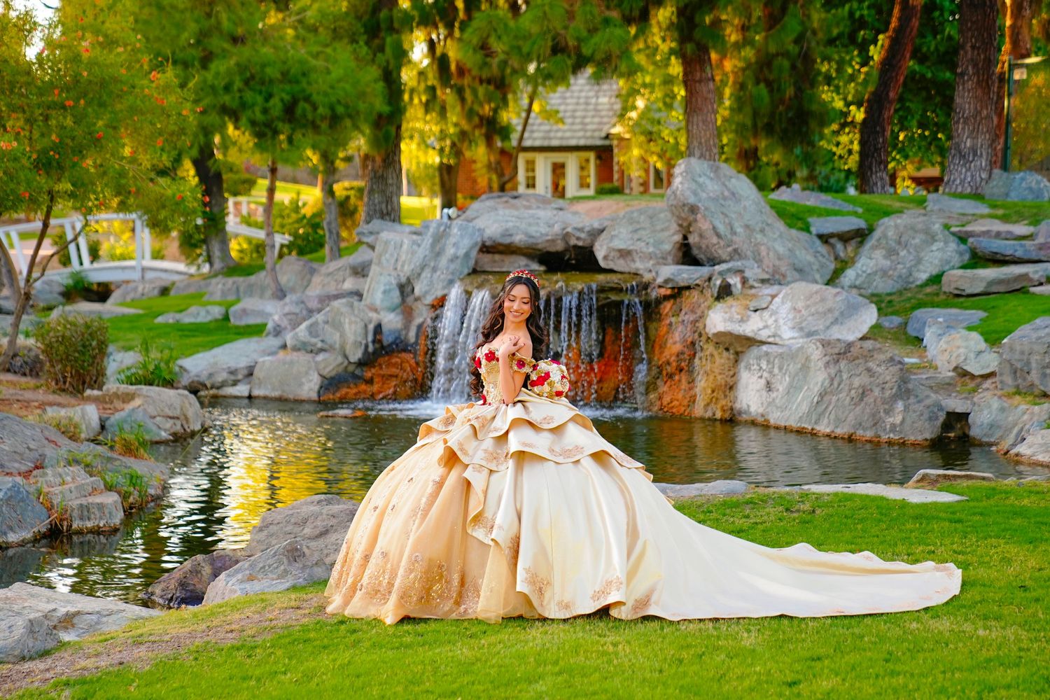 Young woman in a gold quinceañera ball gown posing near a scenic waterfall and pond in a garden.
