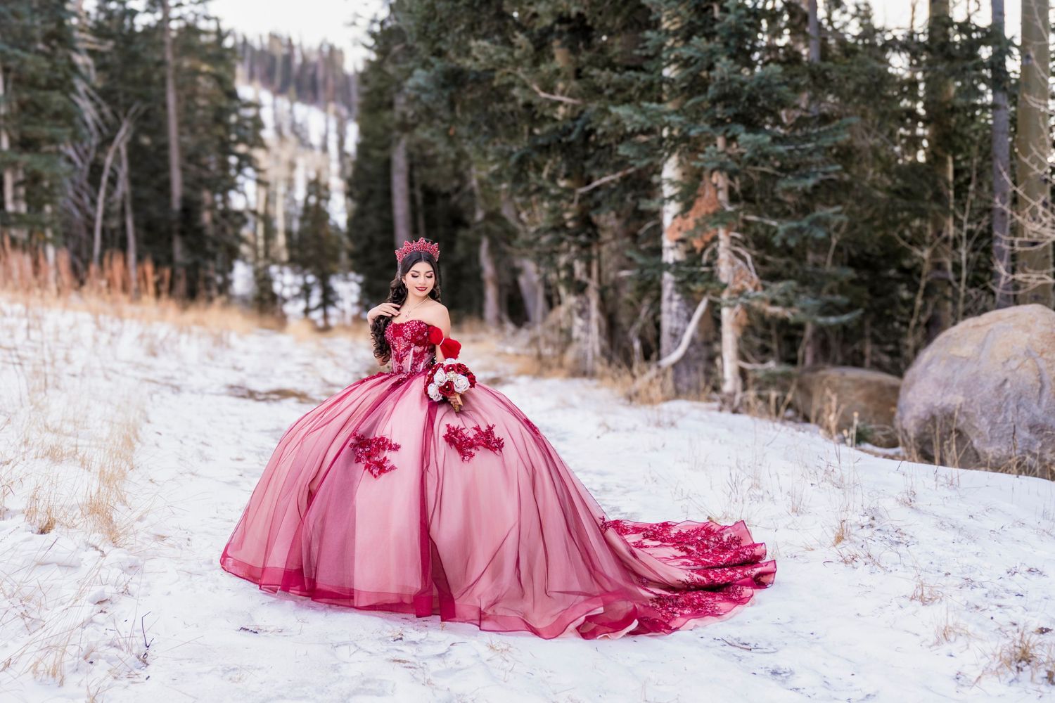 Young woman in a burgundy and pink ball gown with floral details poses in a beautiful snowy mountain forest.