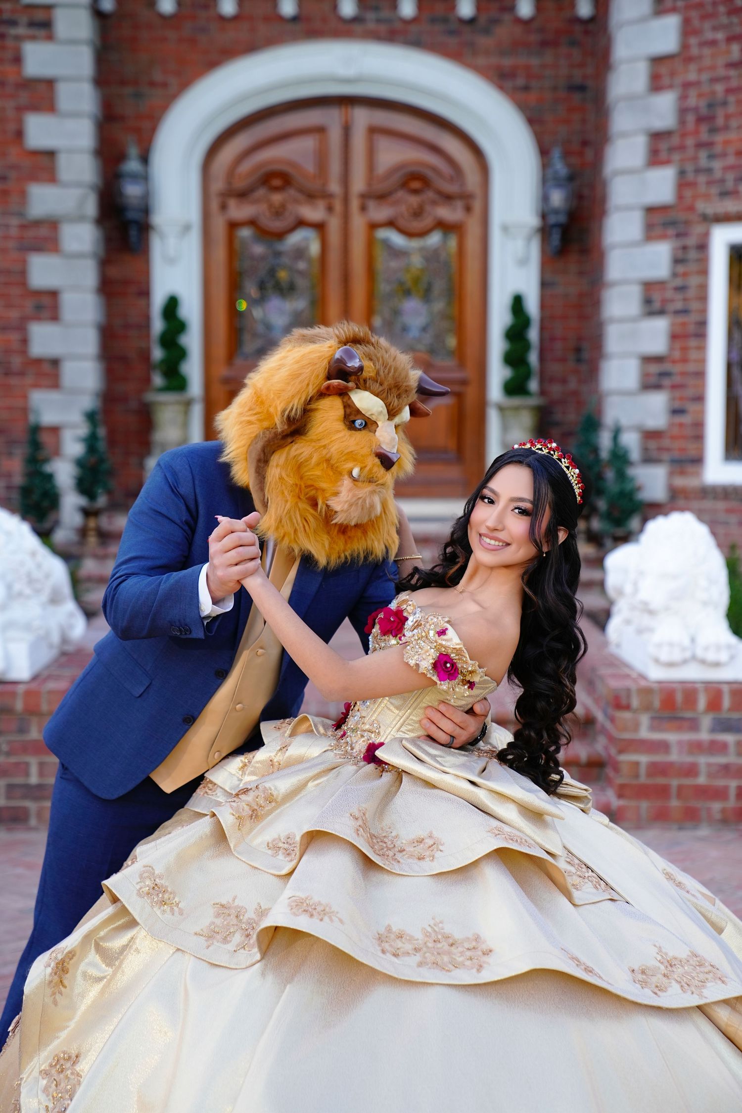 Young woman in gold quinceañera gown posing with Beauty and the Beast character outside brick mansion.