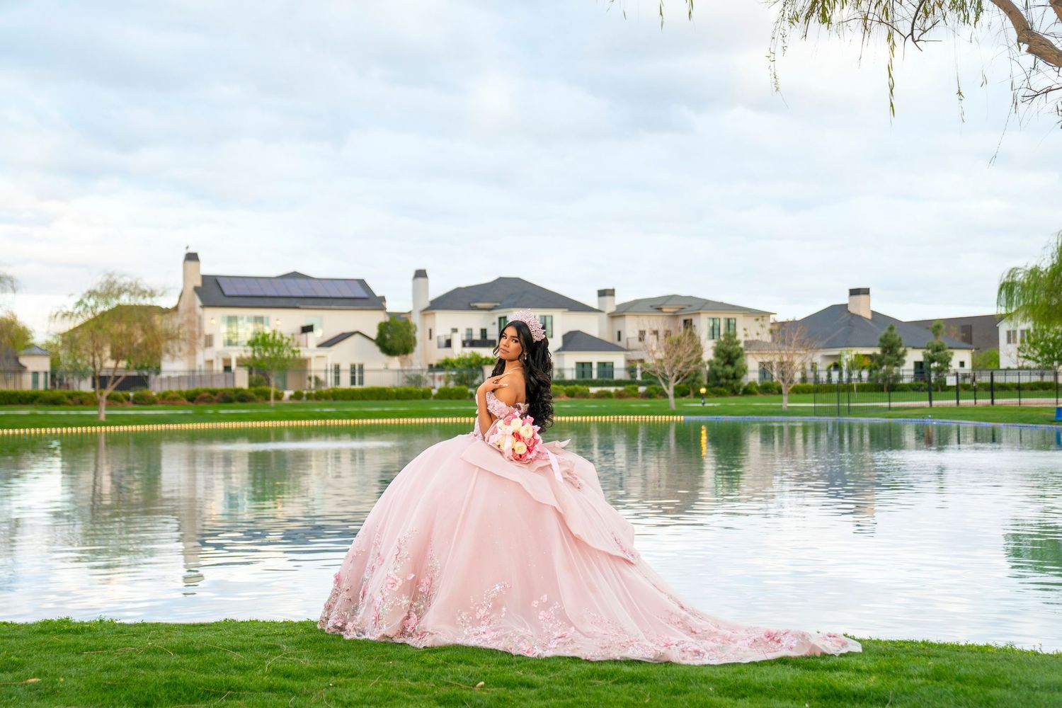 Young woman in a pink ball gown quinceañera dress poses by a serene lake with luxury homes in the background.