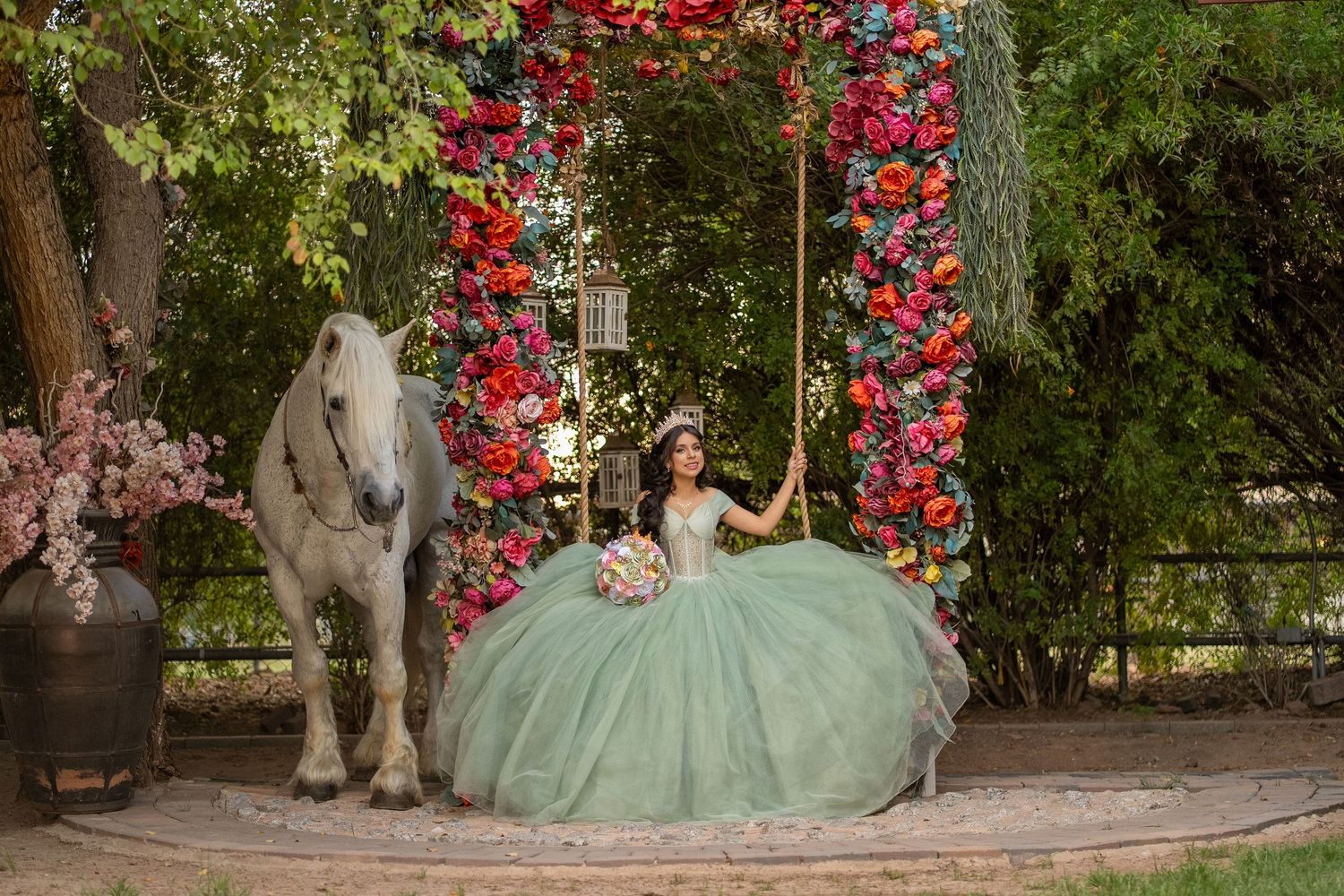 A young woman in a sage green quinceañera gown sits on a floral swing beside a white horse outdoors.