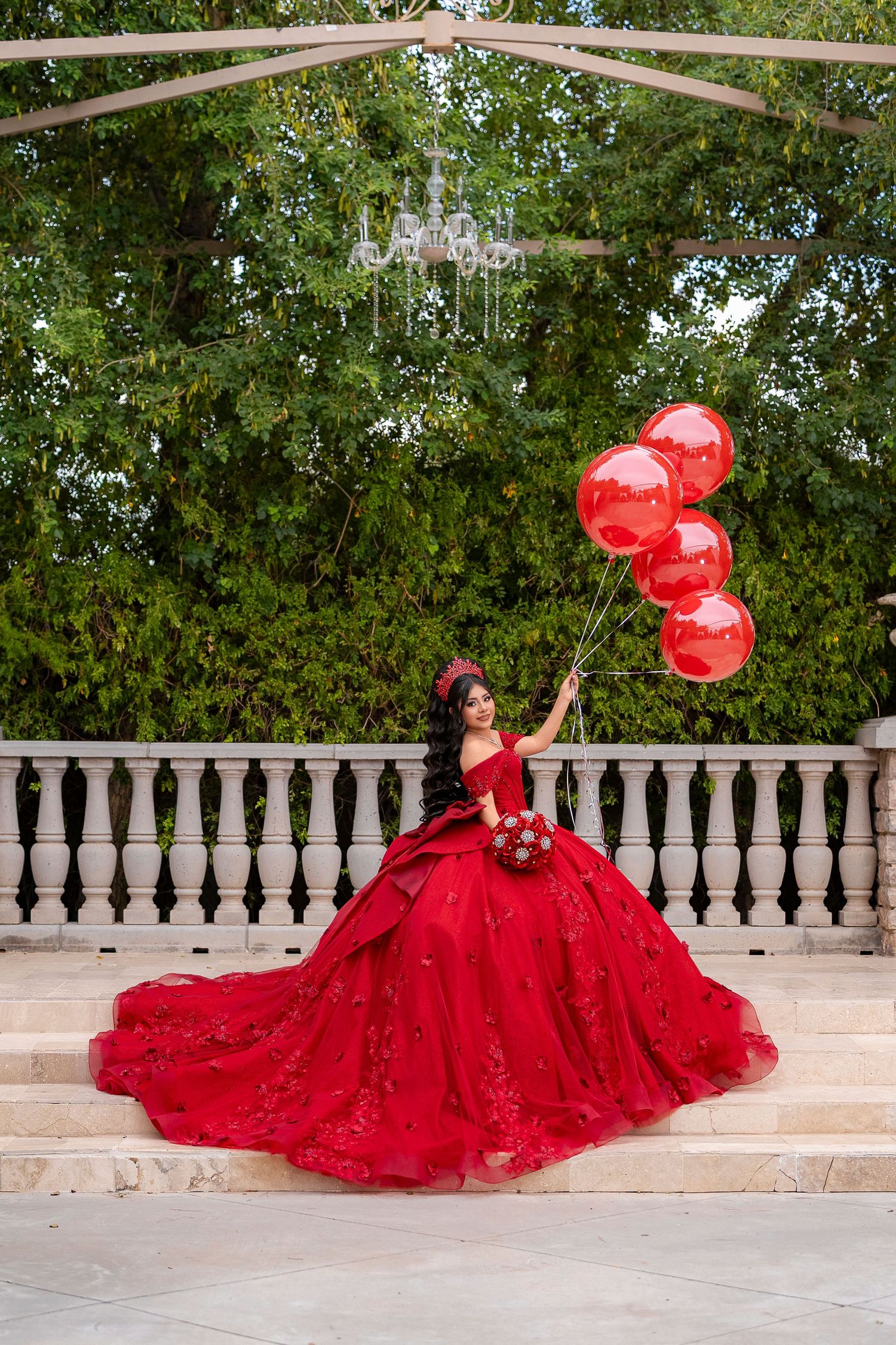 Quinceañera in red ball gown holds red balloons on stone terrace surrounded by lush green garden.