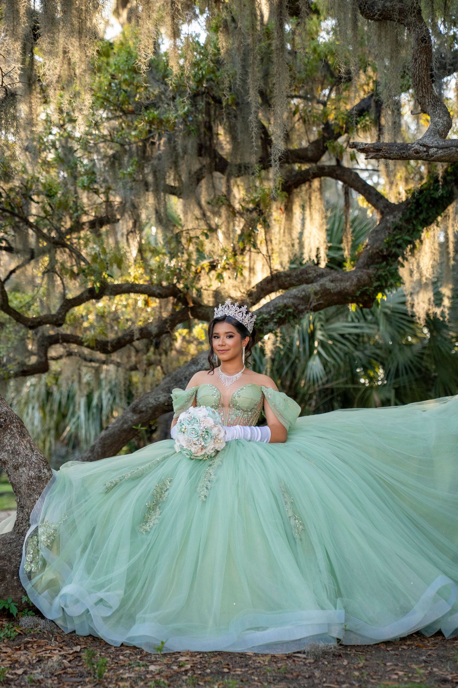 Young woman in mint green quinceañera ball gown with tiara and bouquet poses under moss-draped oak trees.